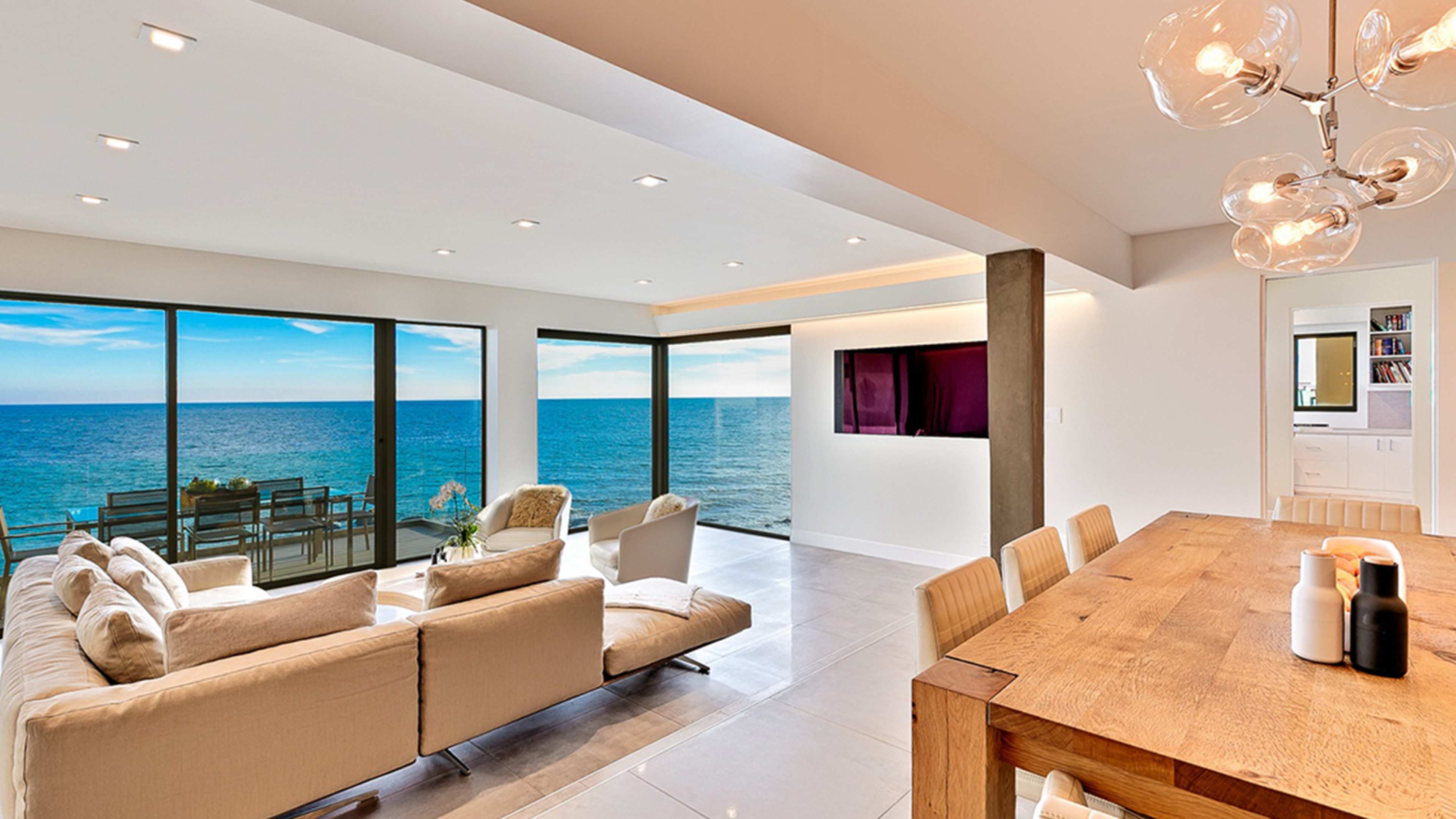 A modern living area features a spacious layout with large windows overlooking the ocean, a comfortable sofa, and a dining table made of wood.