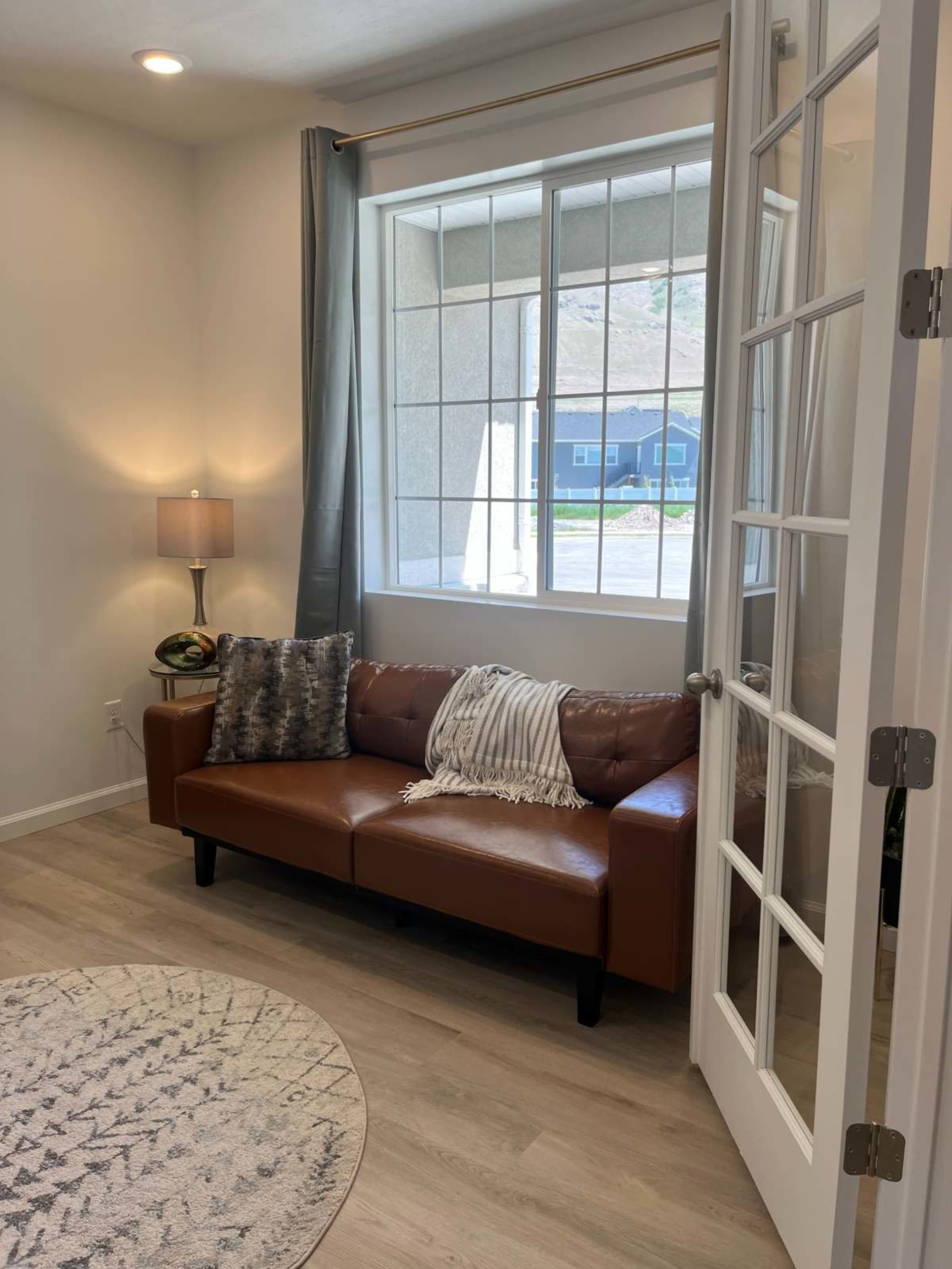 The image shows a well-lit room featuring a brown leather couch, a lamp, and a large window with sheer curtains.