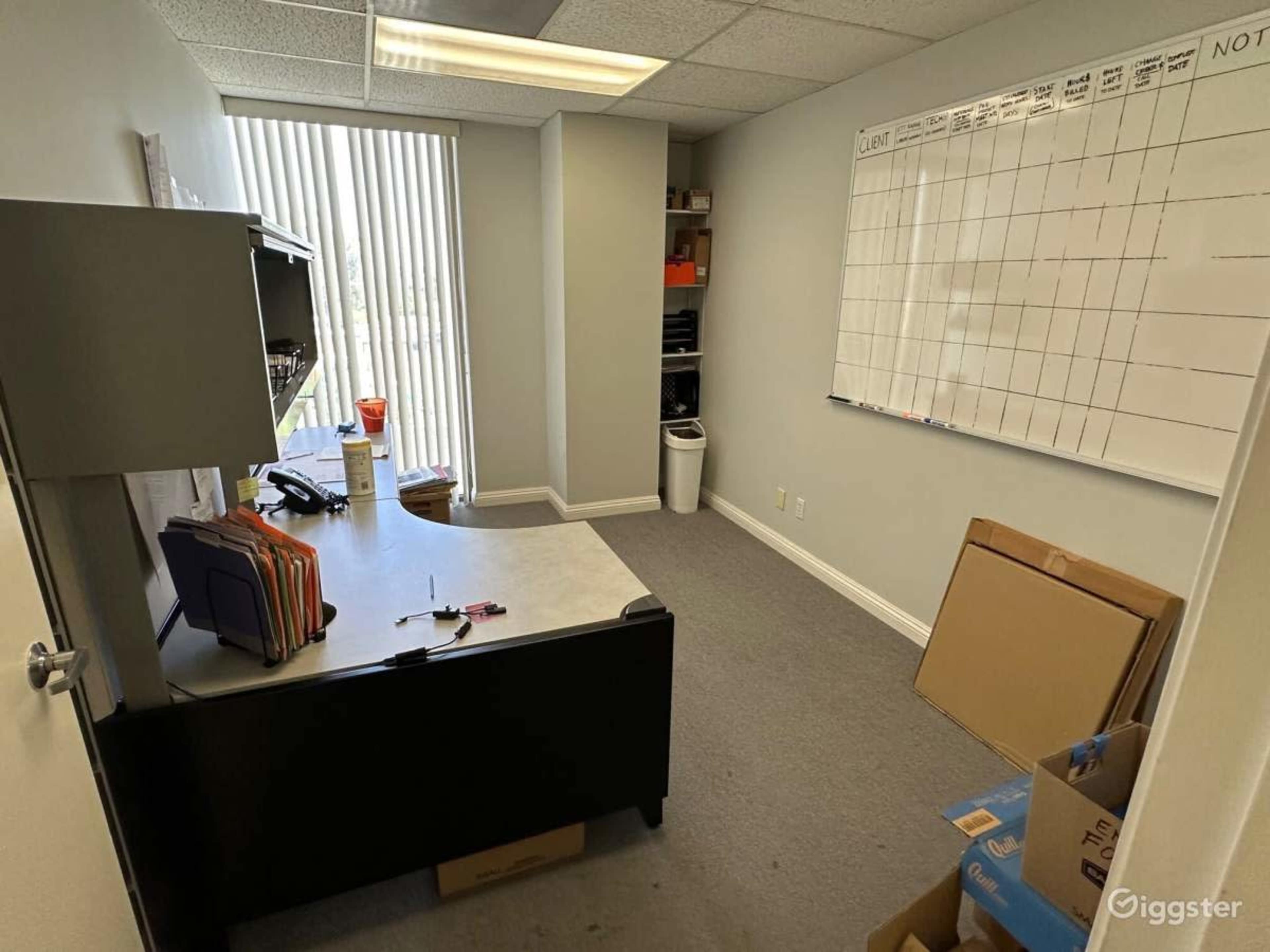 A small office space with a desk, a chair, a whiteboard on the wall, and storage boxes in the corner.