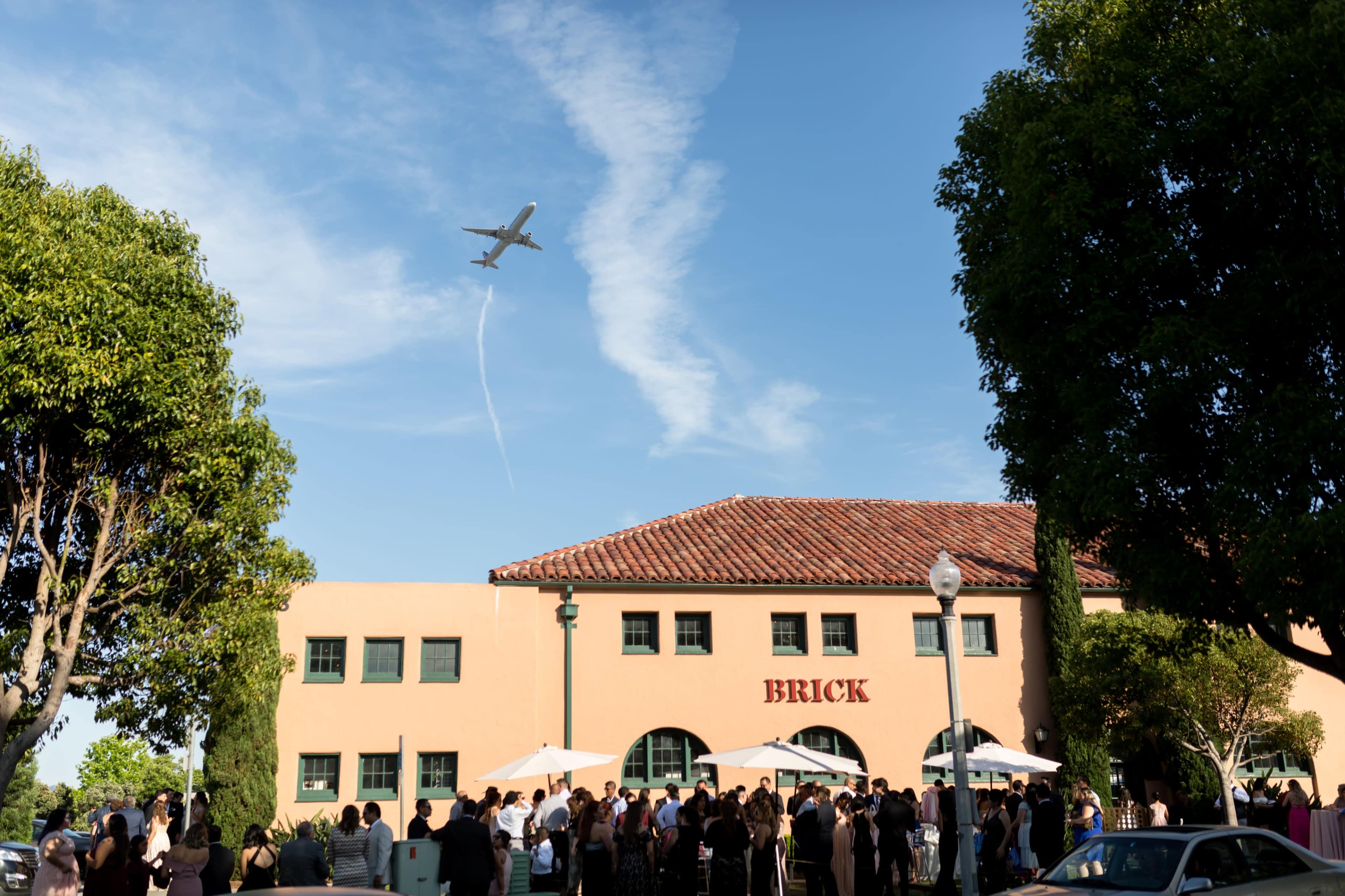 A large crowd gathers outside a building labeled "BRICK" as an airplane flies overhead, leaving a trail in the sky.