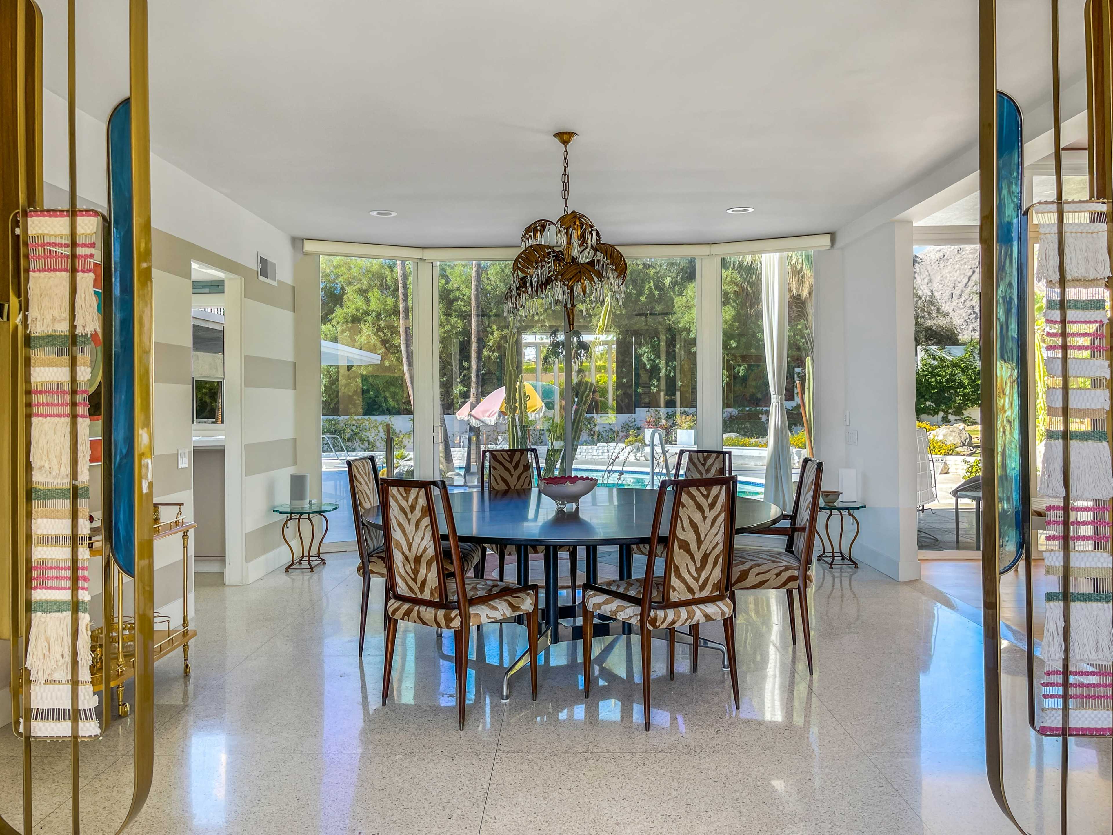 A dining area with a large round table surrounded by patterned chairs, framed by large glass windows that provide a view of a pool and outdoor seating.
