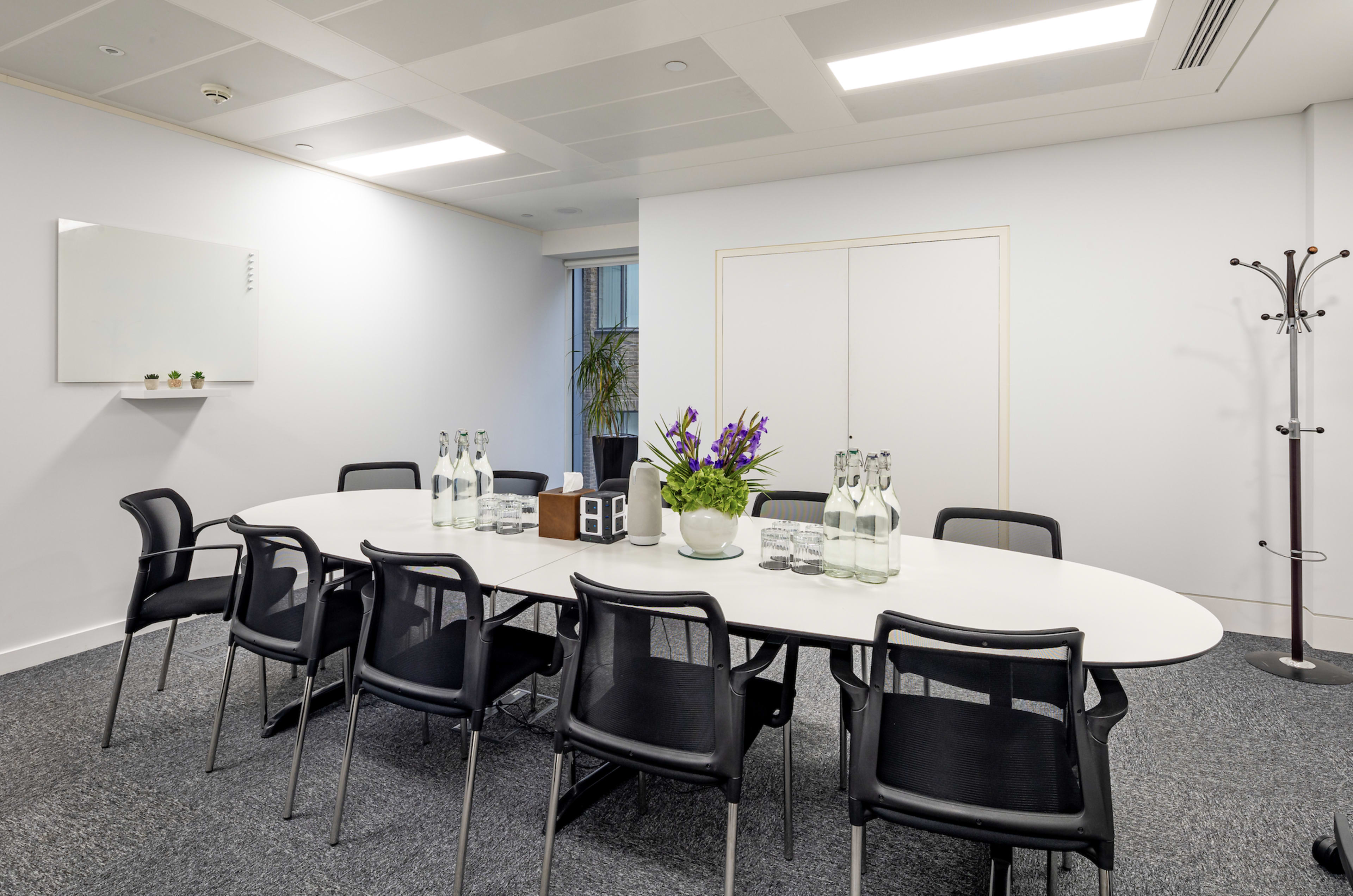 A conference room features a white oval table surrounded by black chairs, with glass water bottles and a flower arrangement in the center.