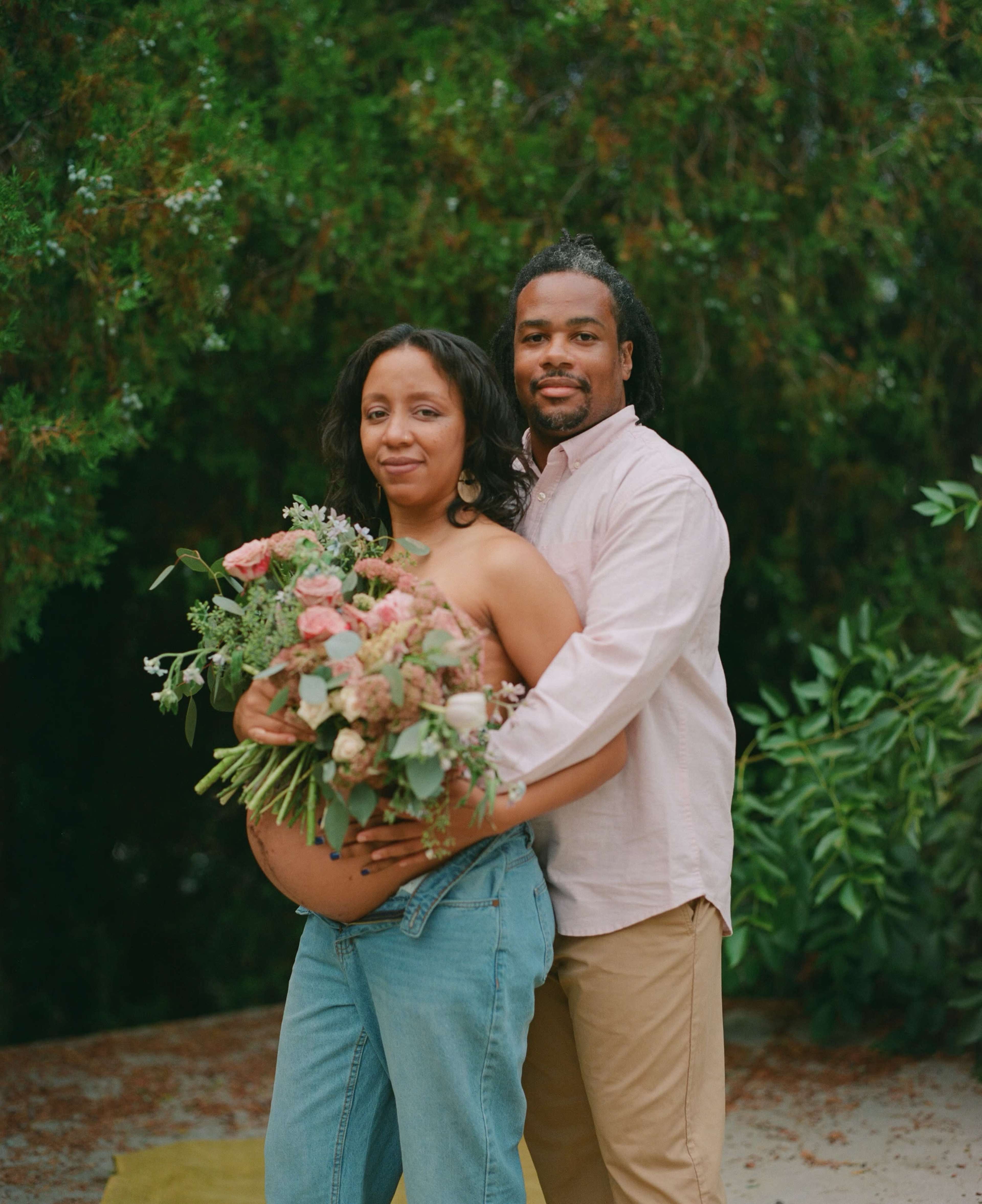 A couple poses together outdoors, with one holding a bouquet of flowers and showcasing a baby bump.