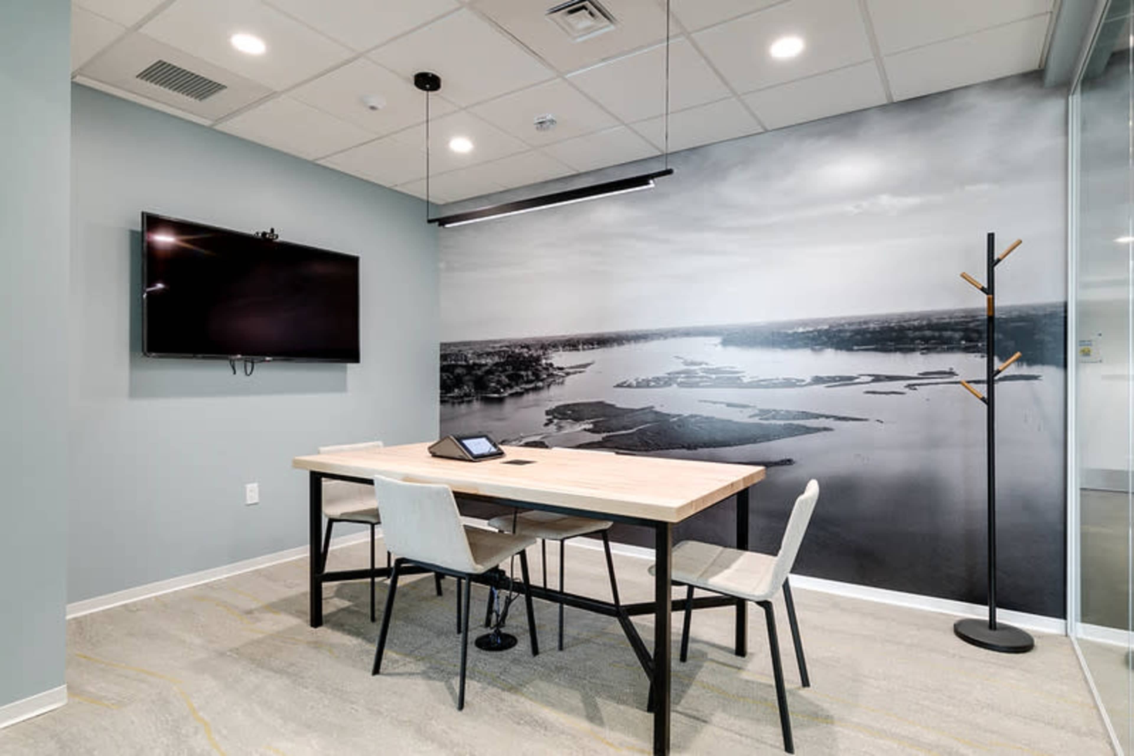 The image shows a modern conference room featuring a large wall mural of a landscape, a wooden table with chairs, and a wall-mounted television.