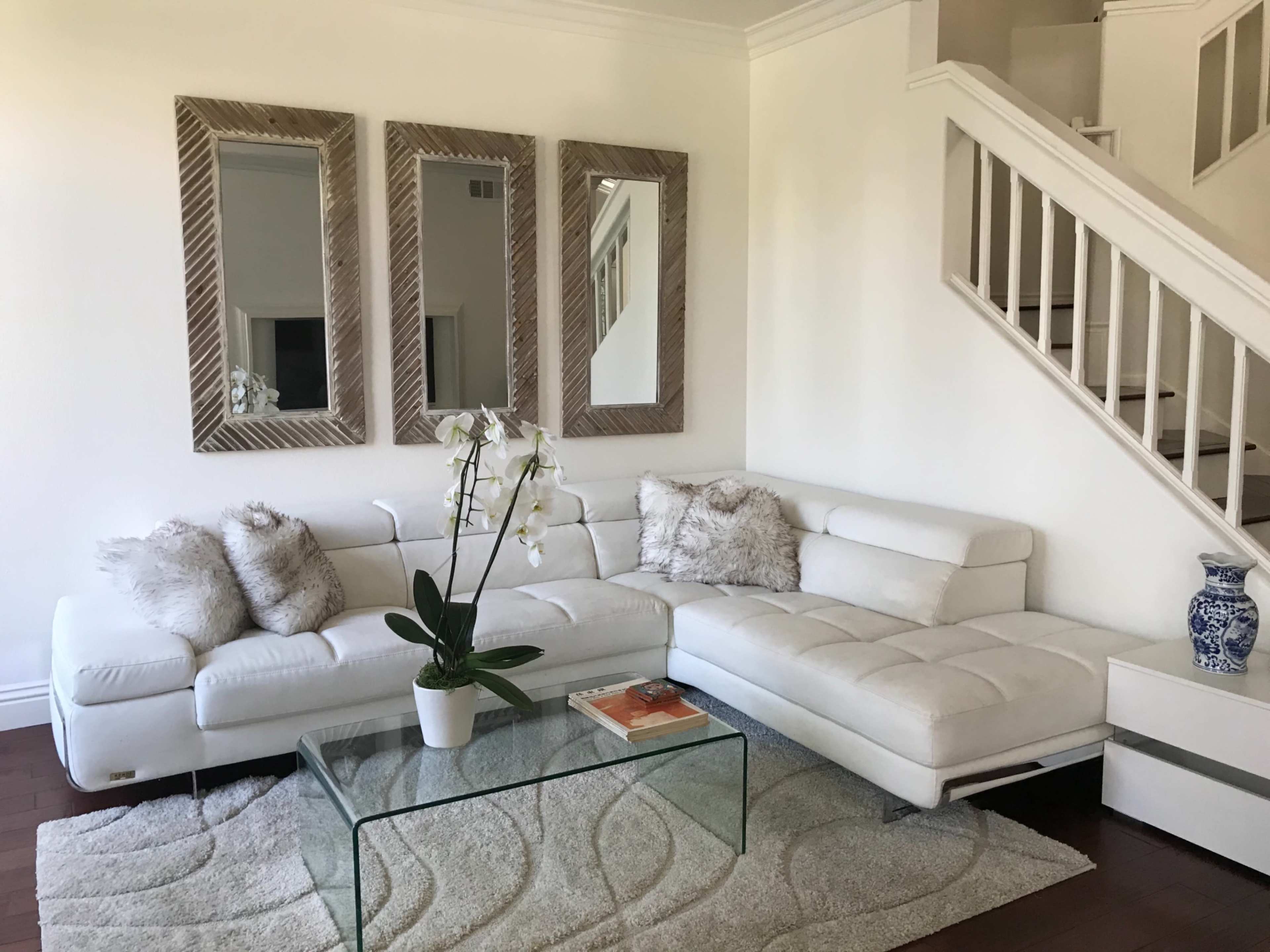 A modern living room with a white sectional sofa, a glass coffee table, and three mirrors mounted on the wall.