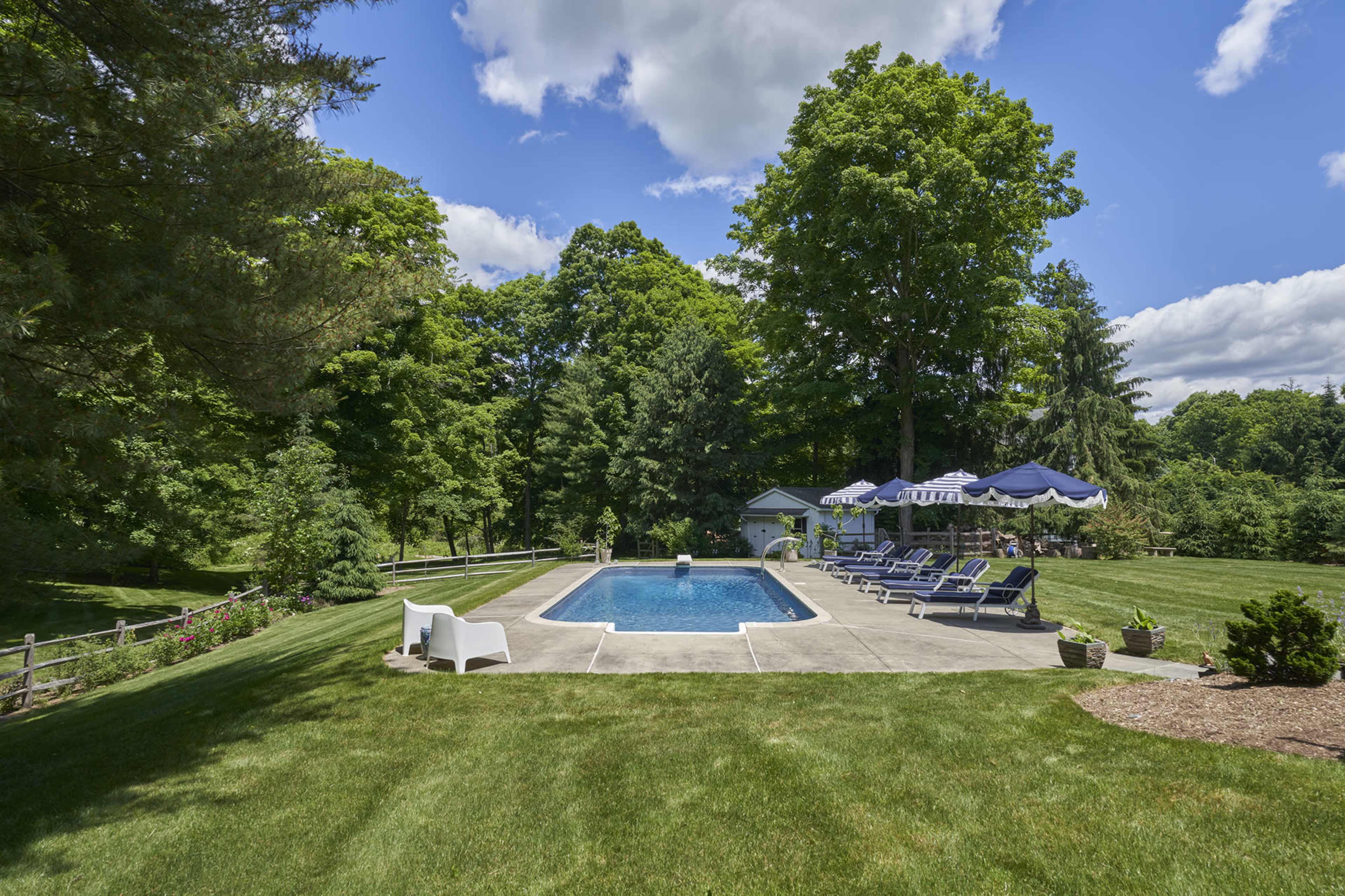 A rectangular swimming pool is surrounded by green grass and various lounge chairs, with trees and a small structure in the background.