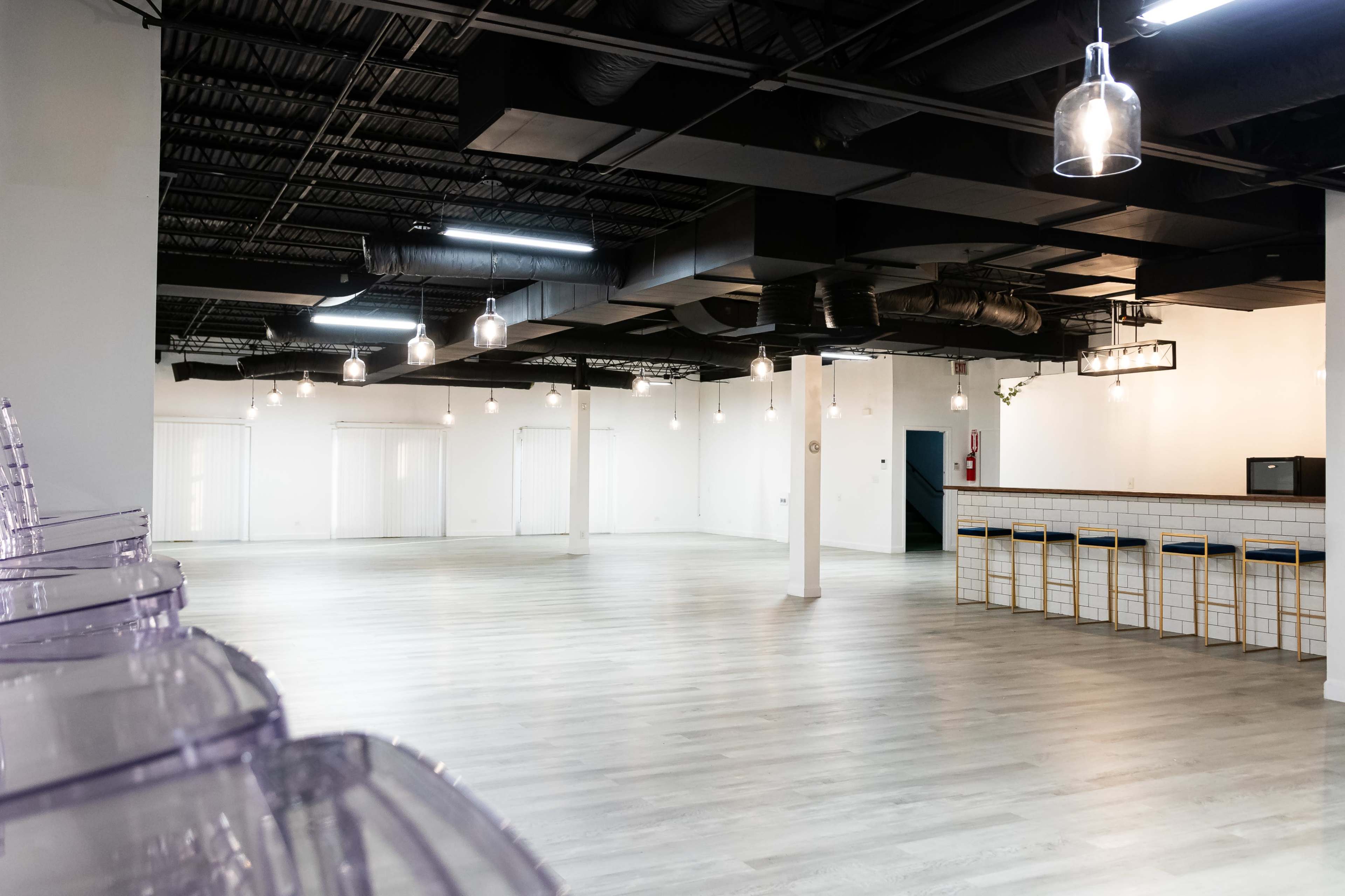The image shows an empty, spacious room with light wood flooring, overhead pendant lights, and a bar area at one end.