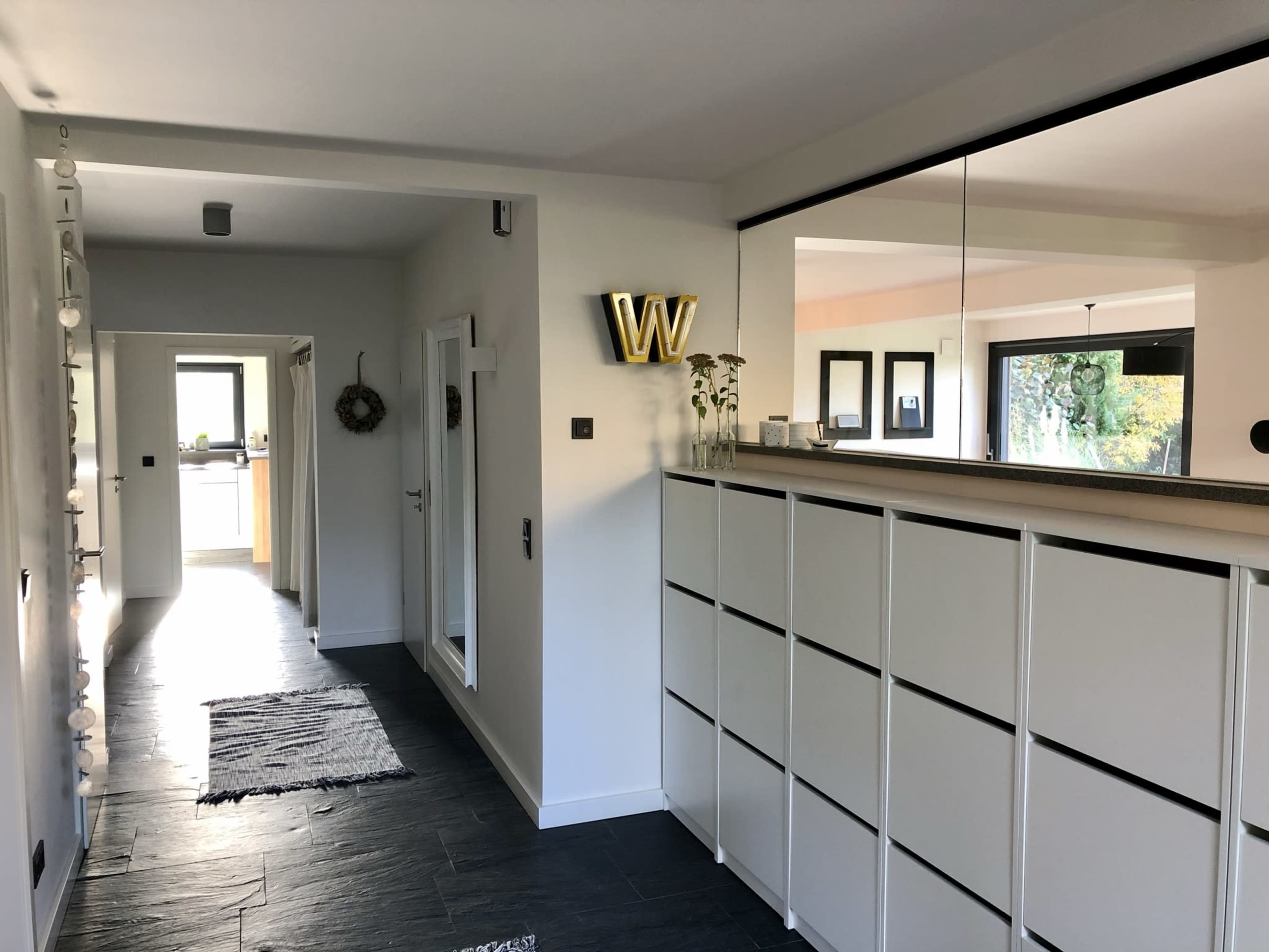 The image shows a modern hallway with a white cabinet along one wall, a reflection in a mirror, and a visible entrance to a kitchen area in the background.