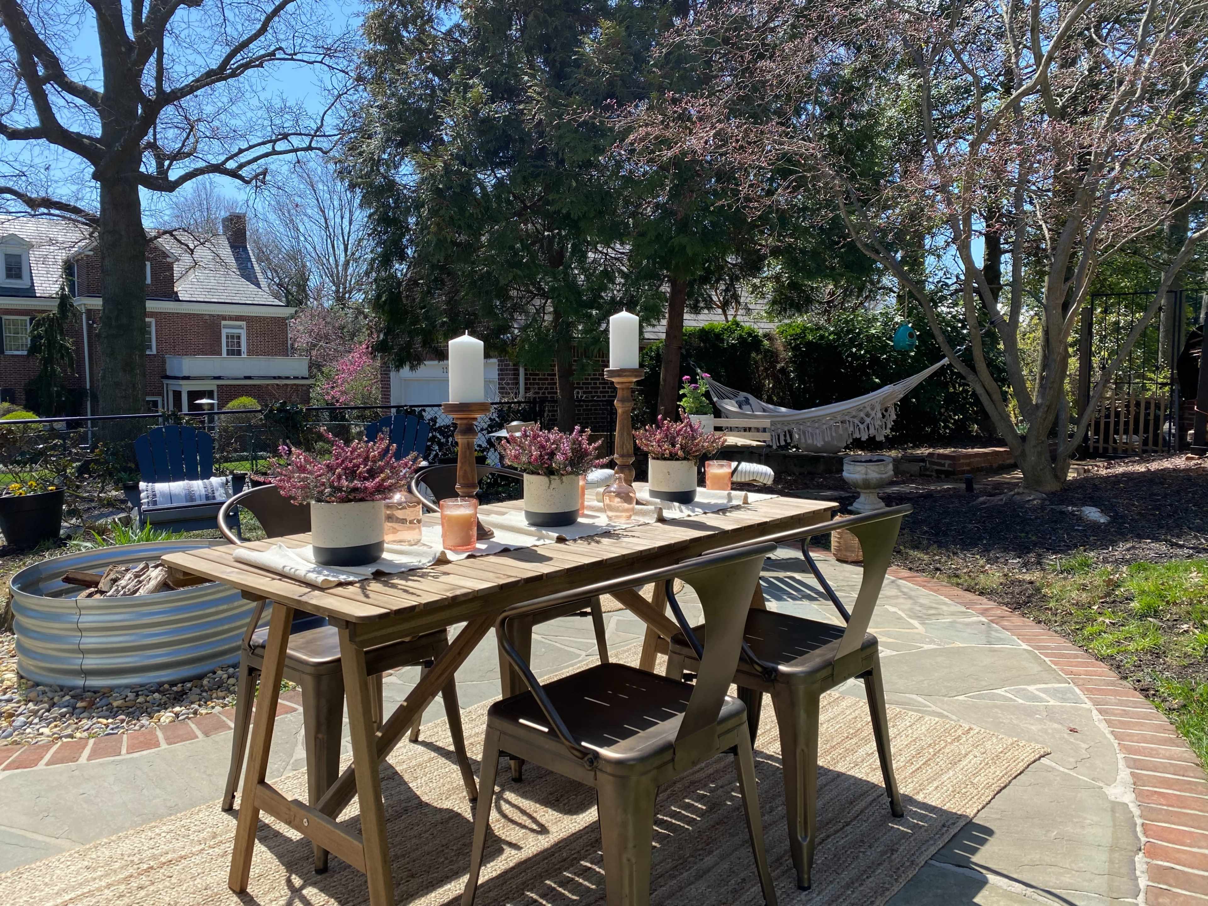 A wooden outdoor dining table is set with candles and plants, surrounded by chairs, a hammock in the background, and a landscaped garden.