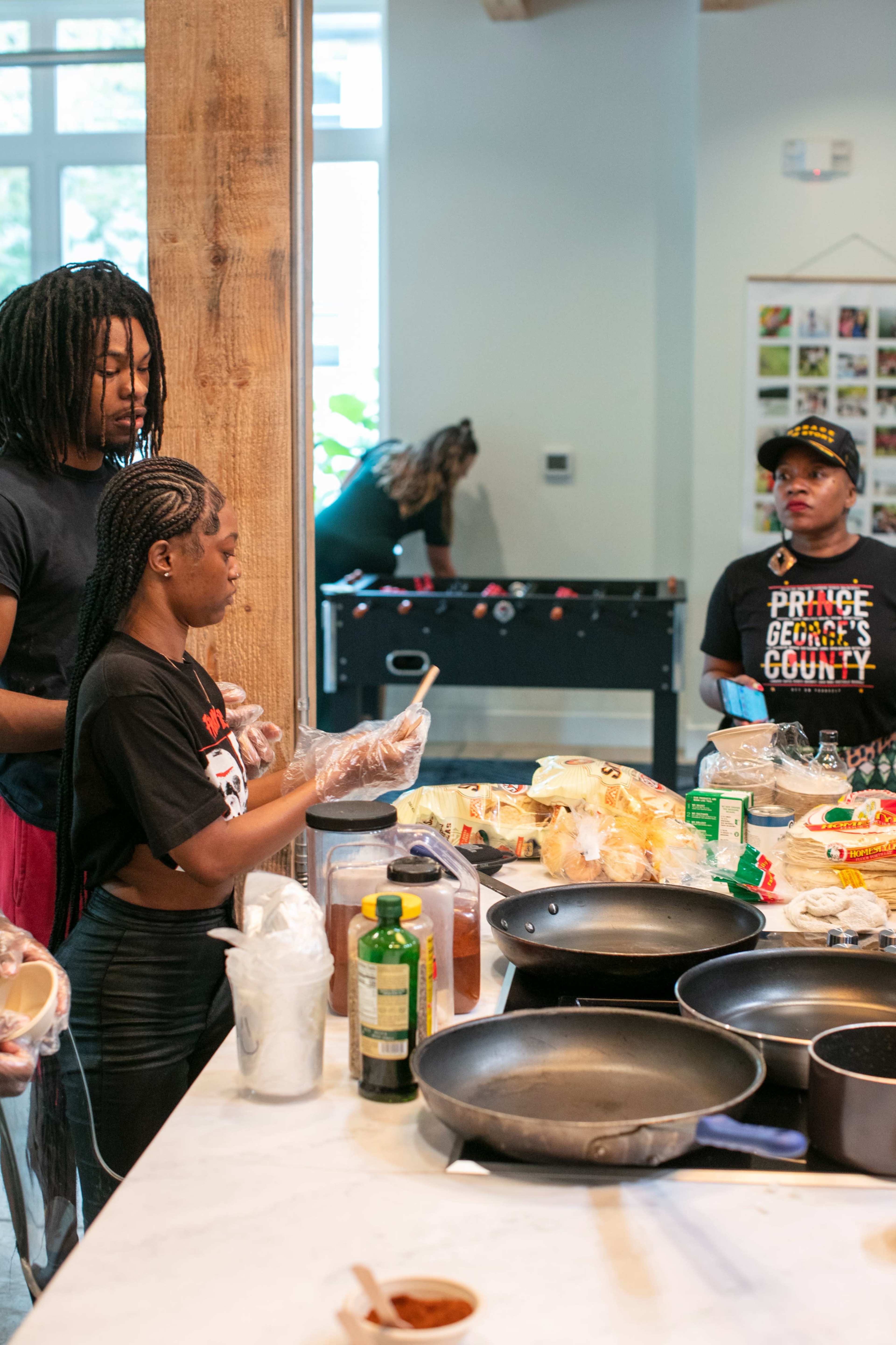 Two individuals are preparing food at a kitchen counter, while another person observes from a distance near a foosball table.