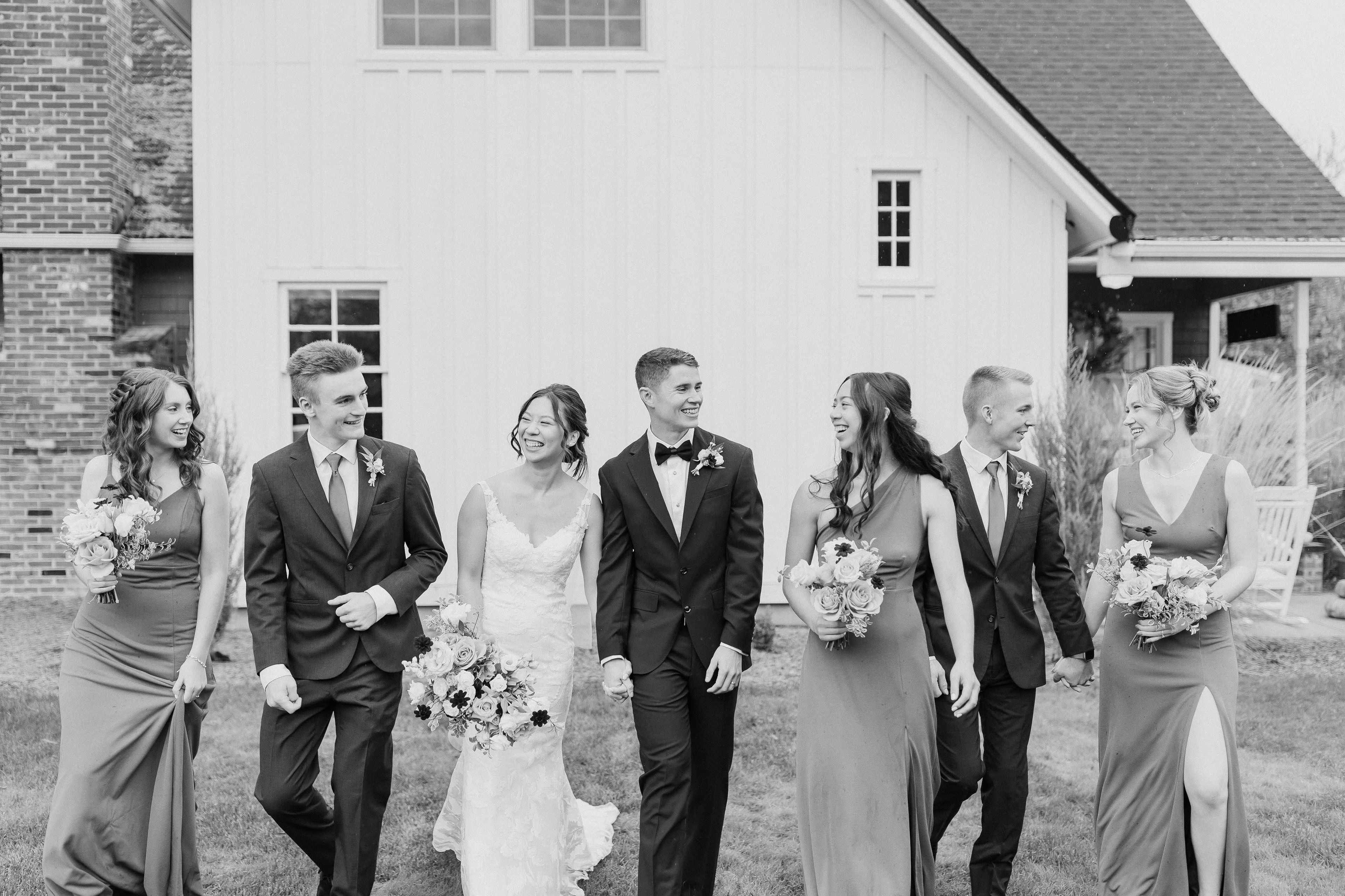A bride and groom walk alongside their wedding party, all smiling and holding floral arrangements, in front of a white-clad building.