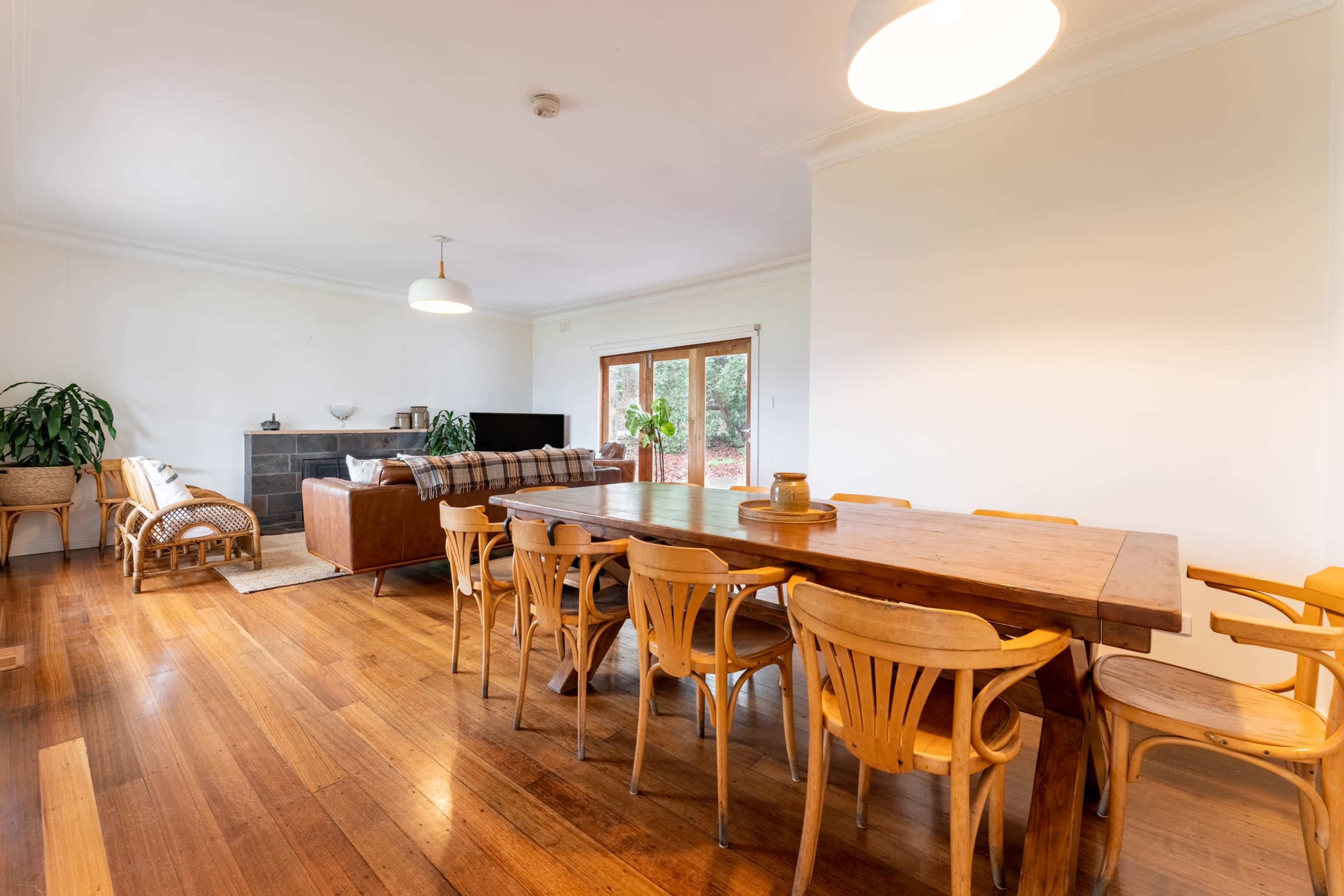 A wooden dining table with multiple chairs is positioned next to a living area featuring a sofa and a large window.