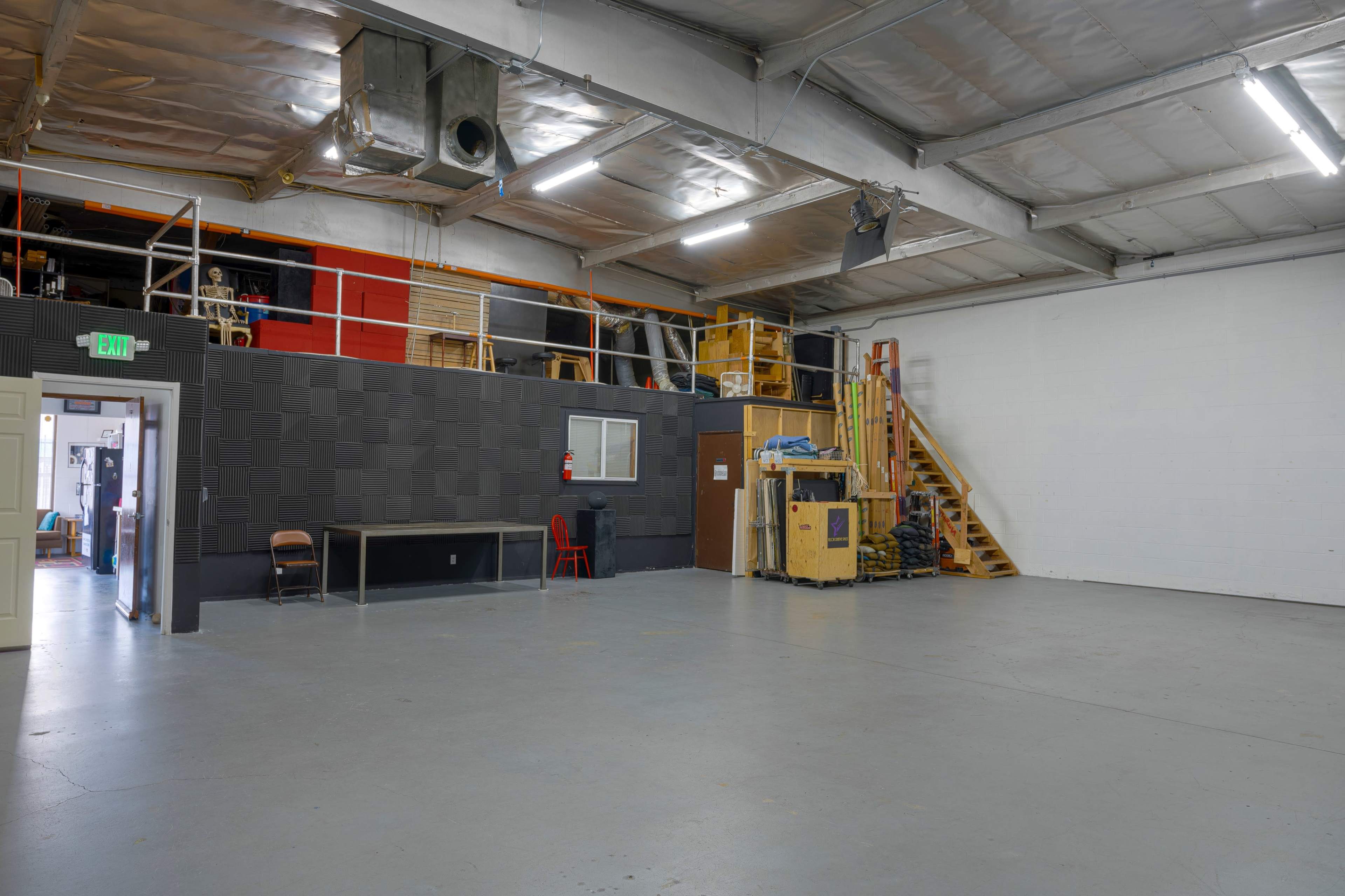 A spacious industrial warehouse with a mix of stacked shelves, a staircase, a table, and various items along the walls.