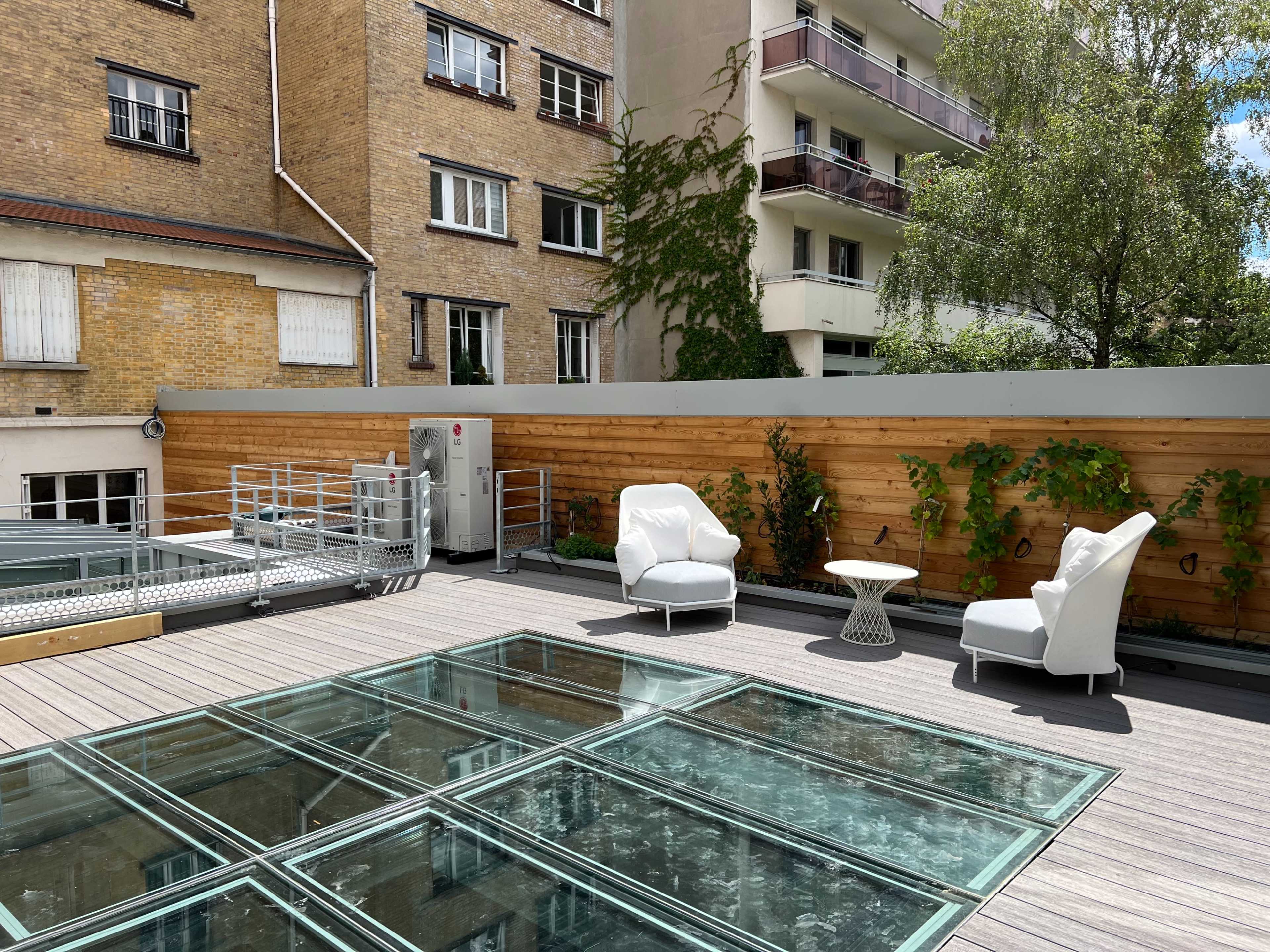 A rooftop terrace with two white armchairs and a small table, surrounded by wooden fencing and green plants.
