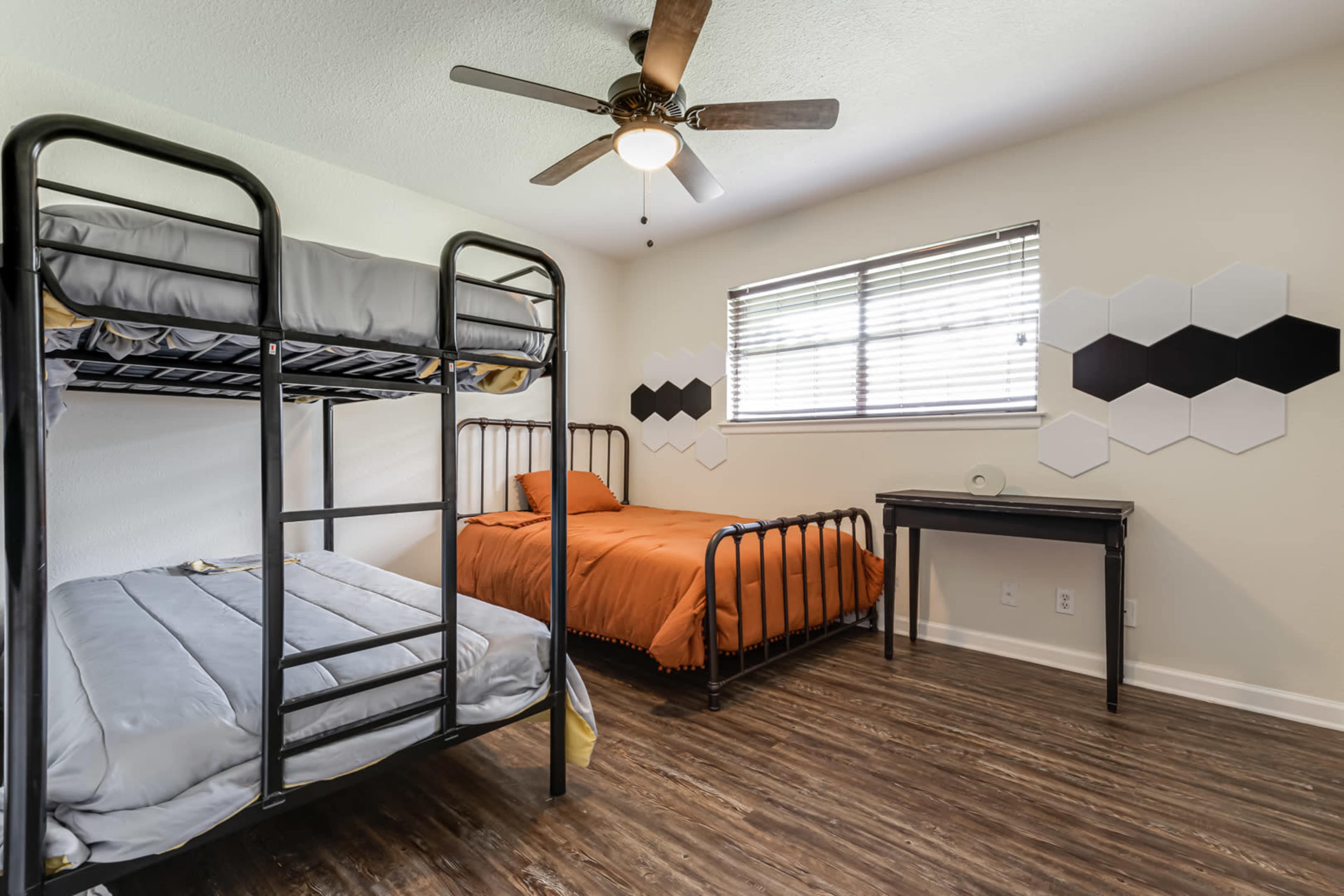 A bedroom with a black metal bunk bed, a separate orange bed, a small desk, and hexagonal wall decor.