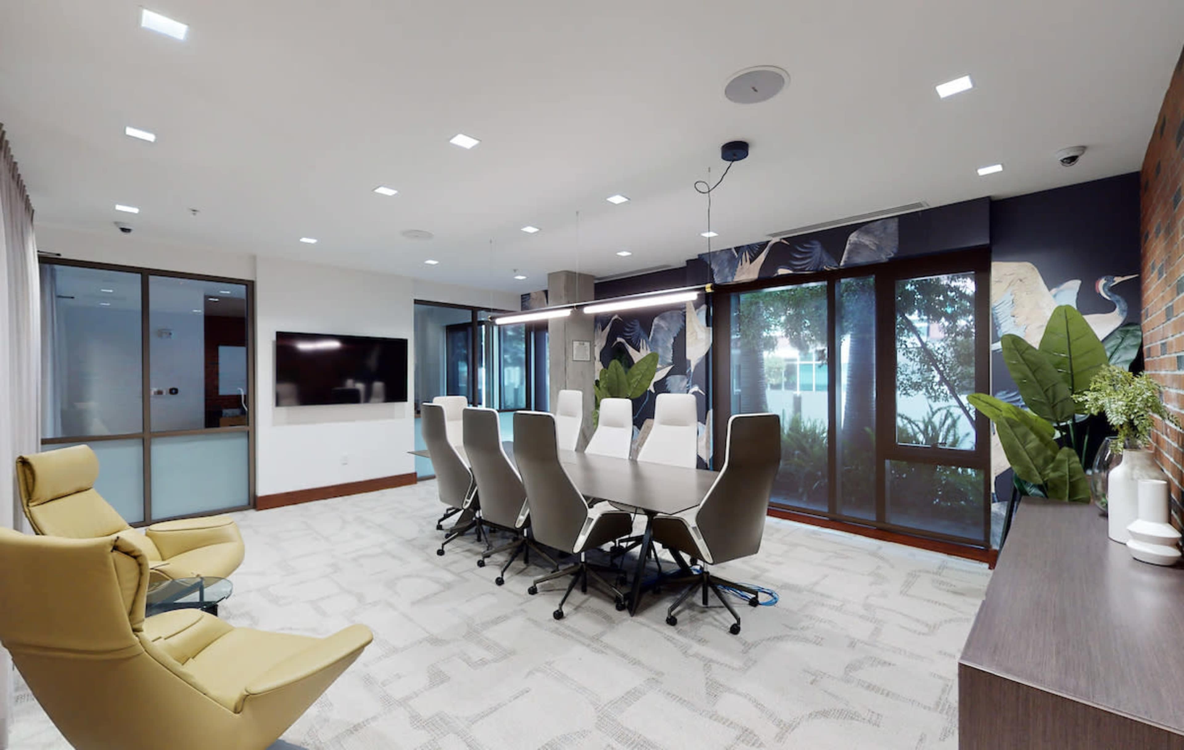 A modern conference room features a long table surrounded by ergonomic chairs, large windows with indoor plants, and a wall-mounted screen.