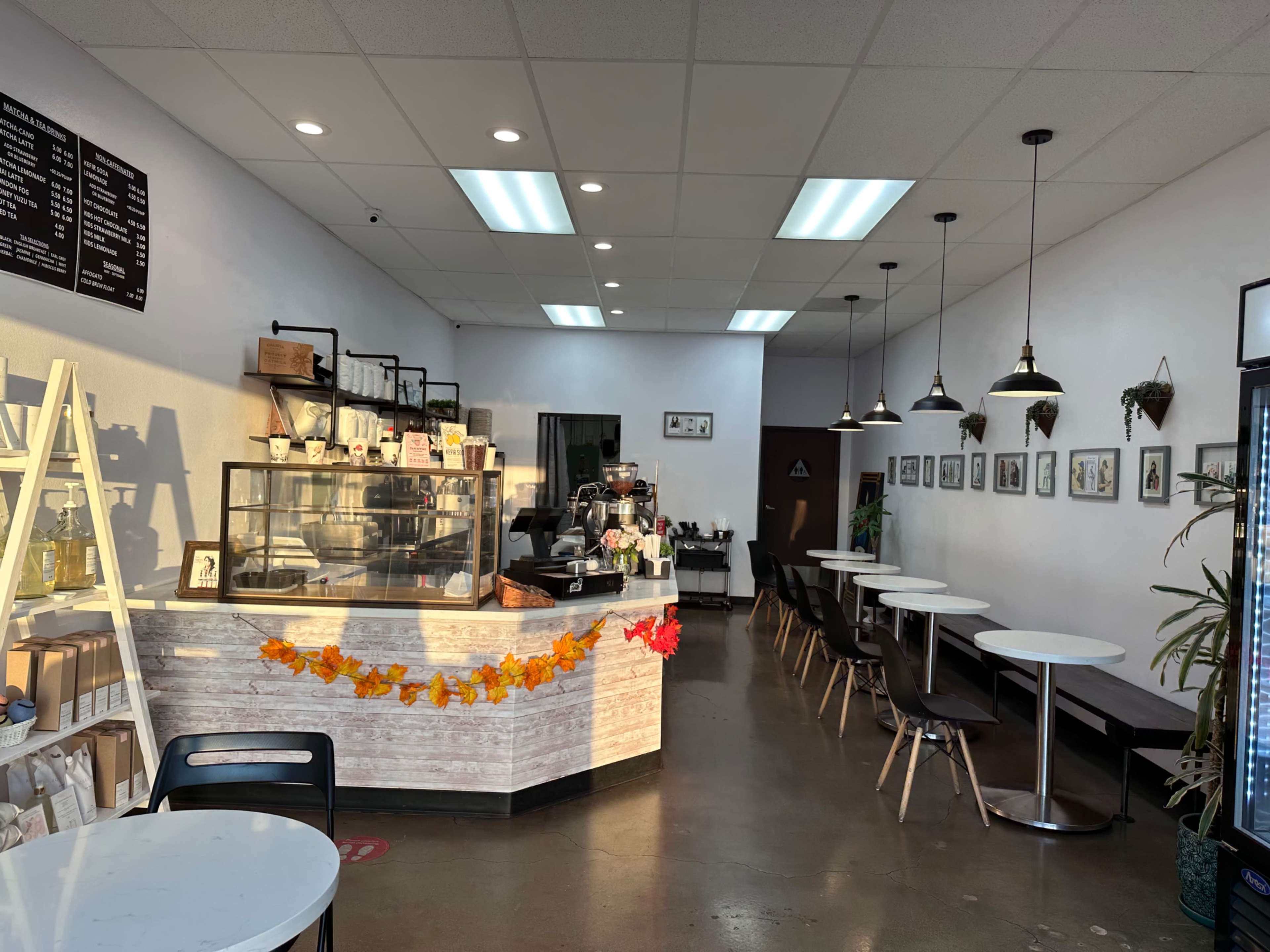 A cozy café interior with a counter displaying beverages, tables arranged along the walls, and framed pictures hanging on the white walls.
