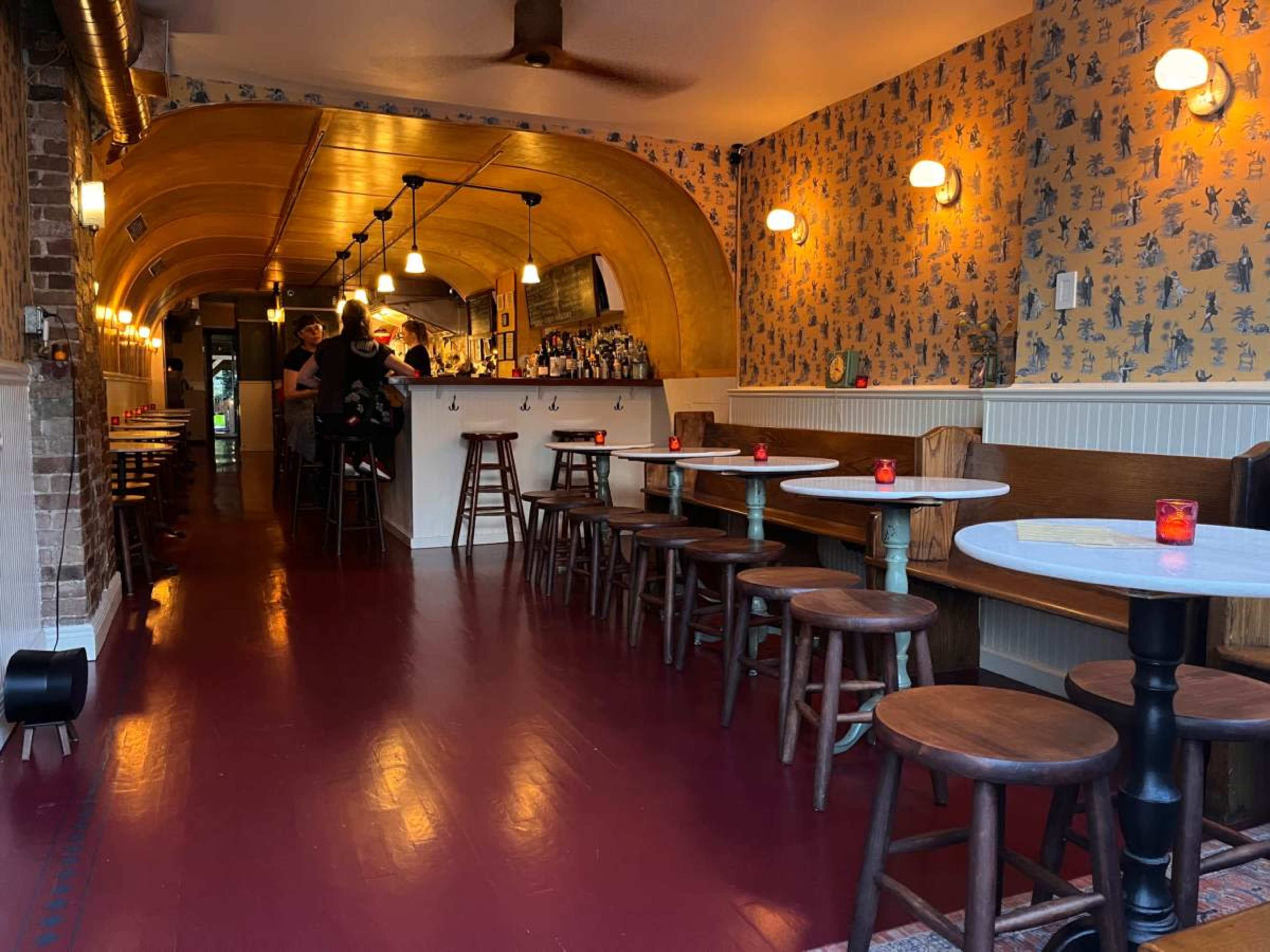 The image shows a narrow restaurant interior with wooden stools and tables, a bar area at the back, and patterned wallpaper along the walls.