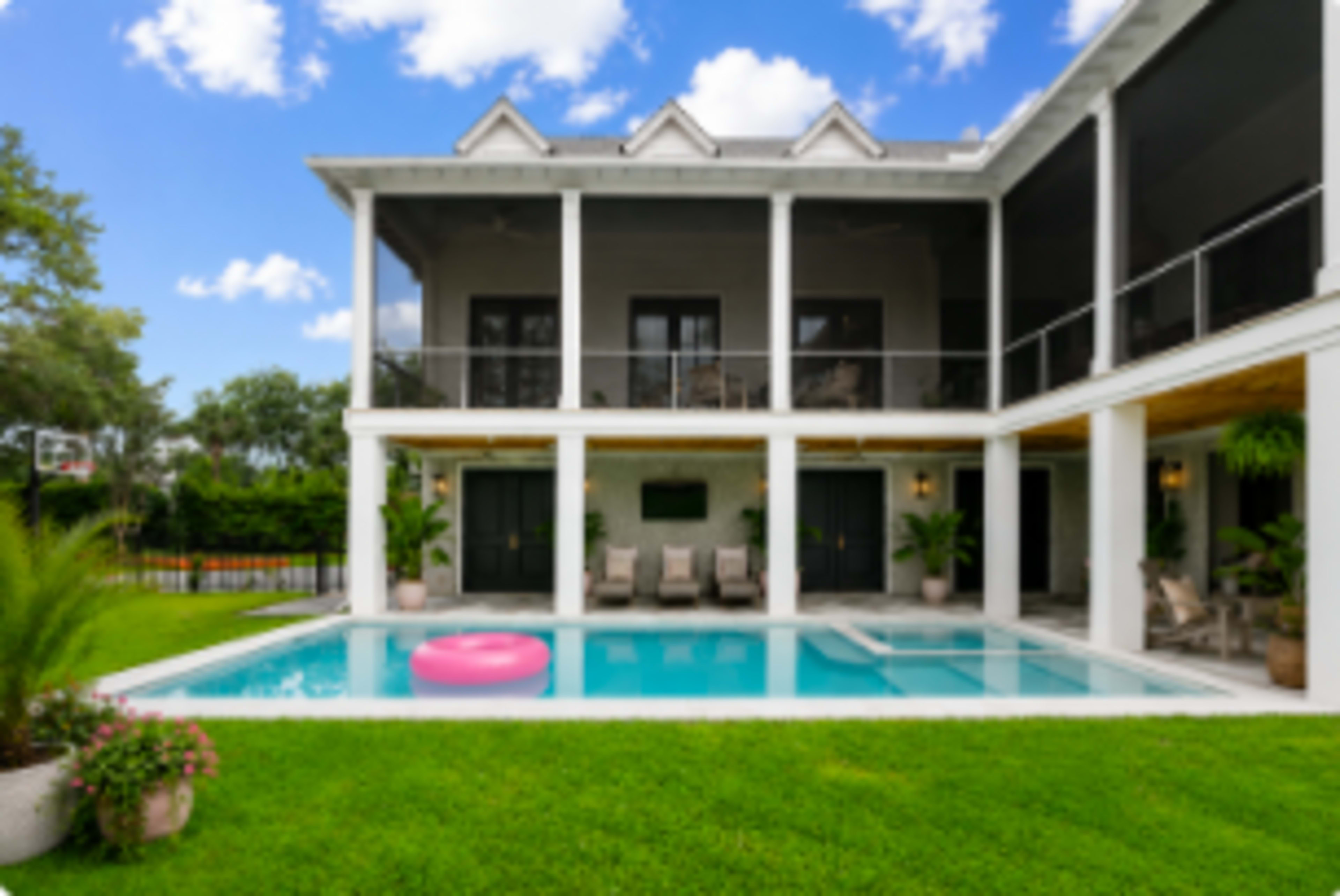 A modern two-story villa features a swimming pool with a pink float, surrounded by a well-manicured lawn and lush greenery.