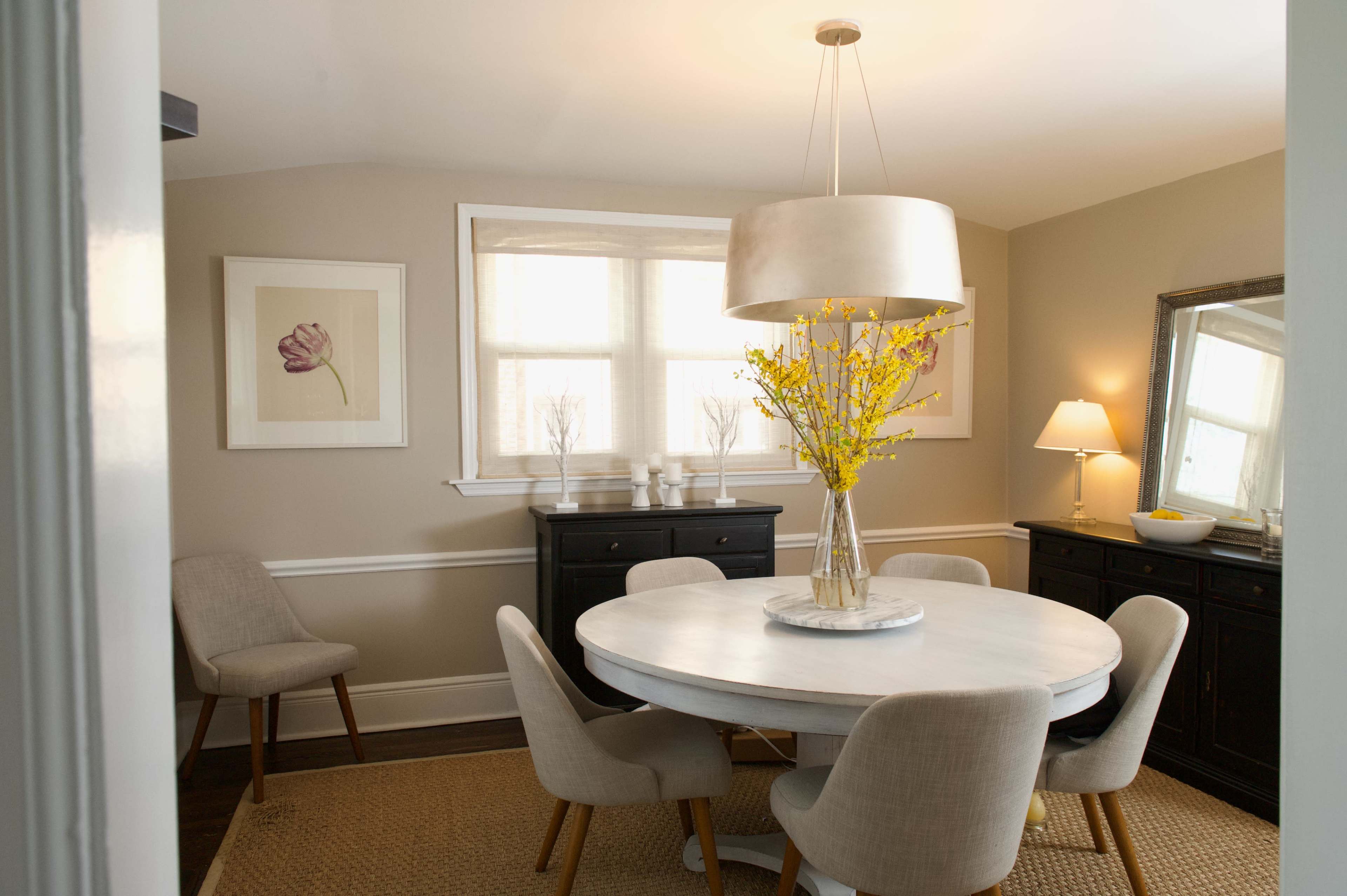 A dining room features a round table surrounded by upholstered chairs, with a large pendant light and a vase of yellow flowers as the centerpiece.