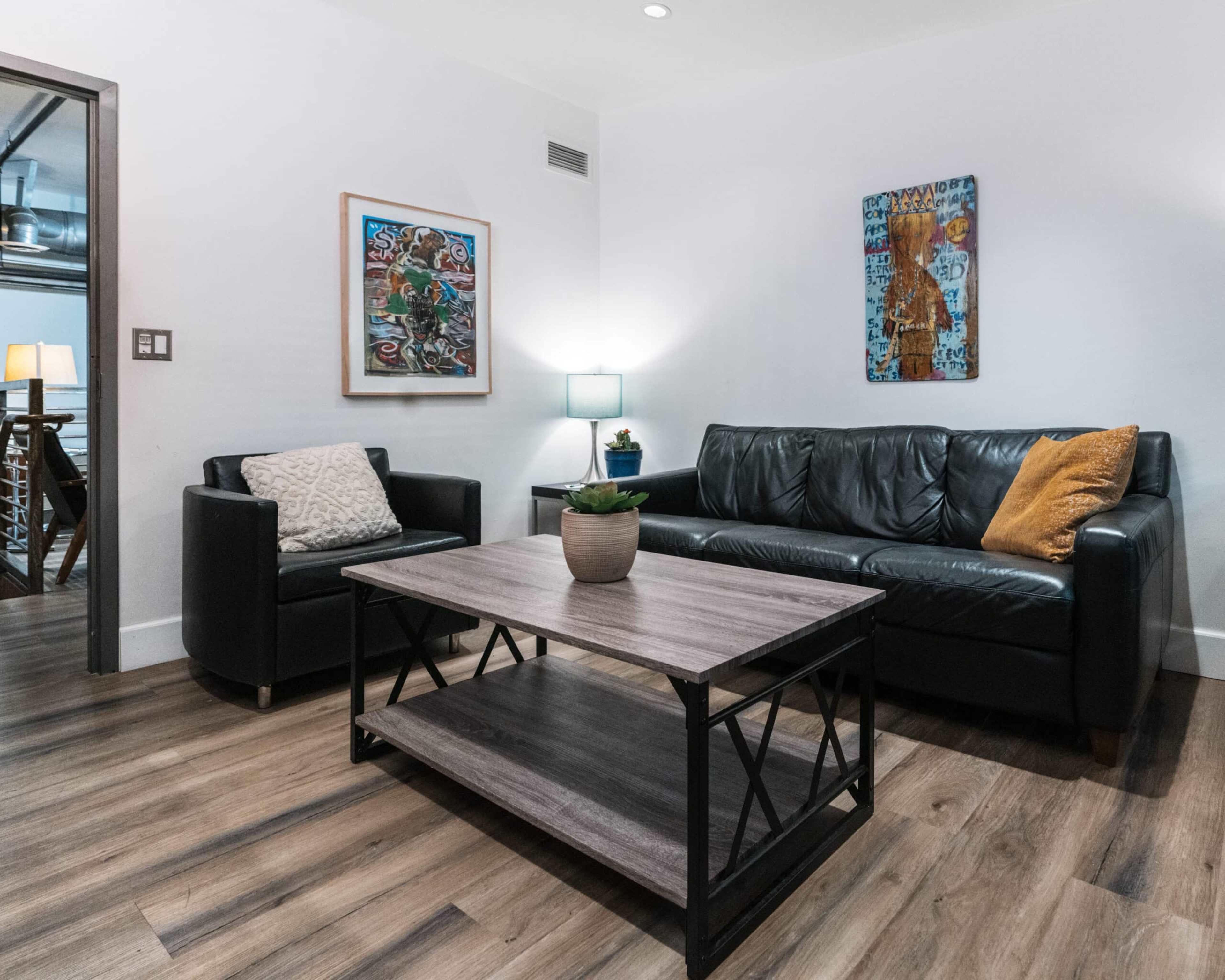 The image shows a modern living room with a black leather sofa, a single armchair, a coffee table, and artwork on the walls.