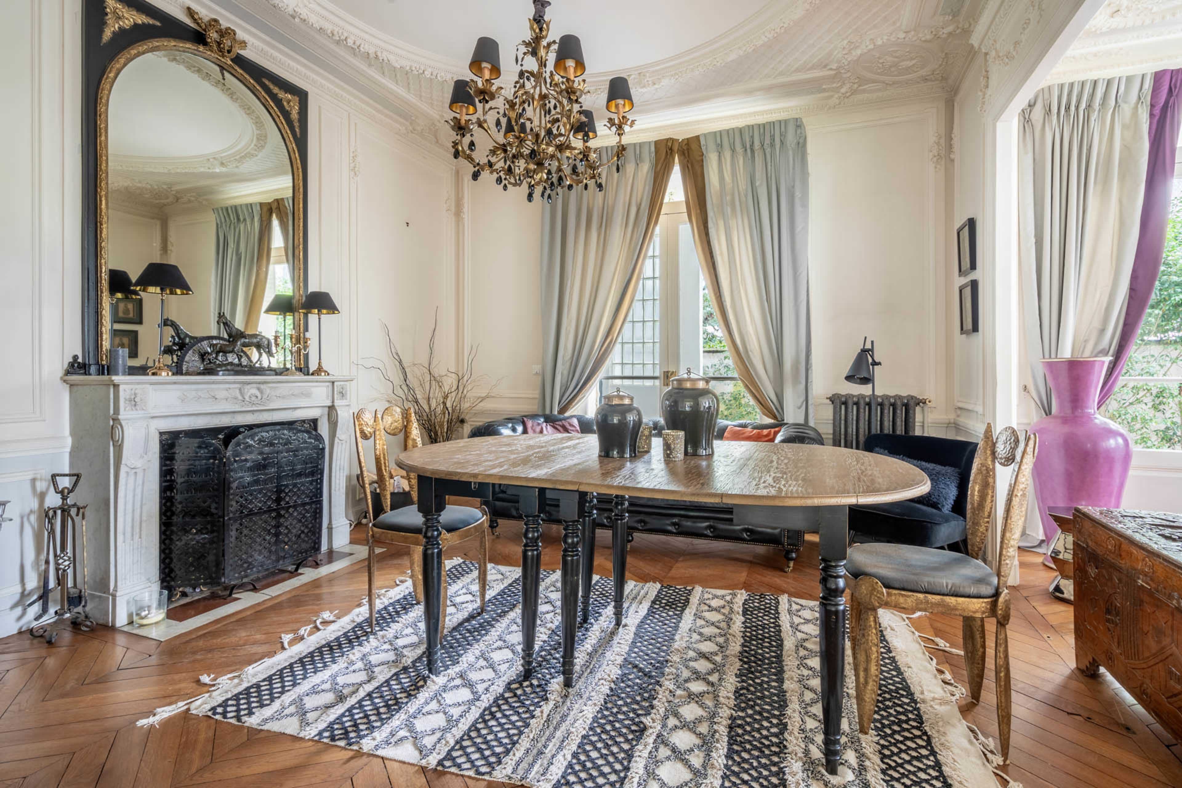 The image shows a decorated dining area with a round table, black chairs, a large mirror, and a chandelier, surrounded by elegant drapes and a patterned rug.