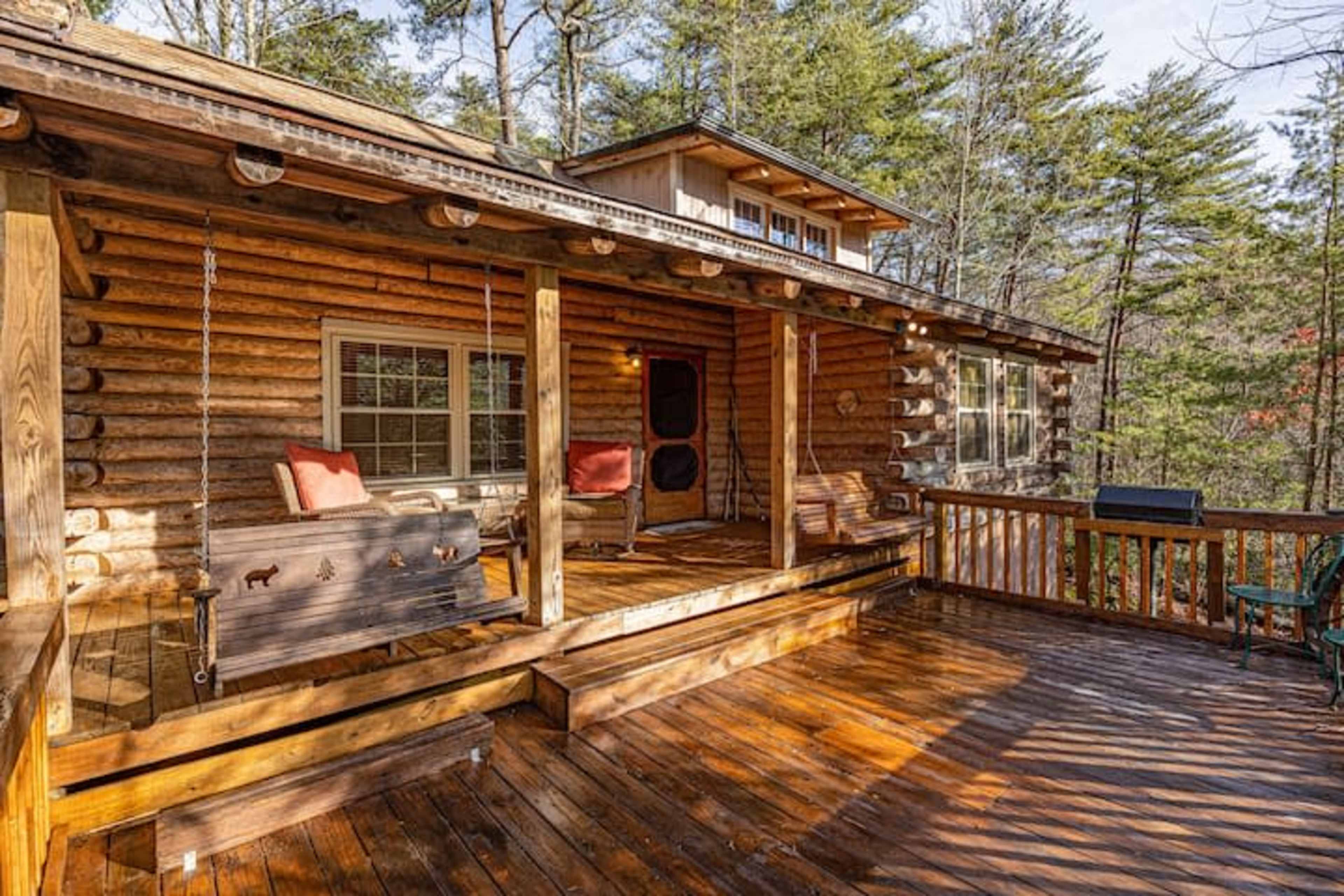 The image shows a wooden log cabin with a front porch featuring a swing and a grill, surrounded by trees.