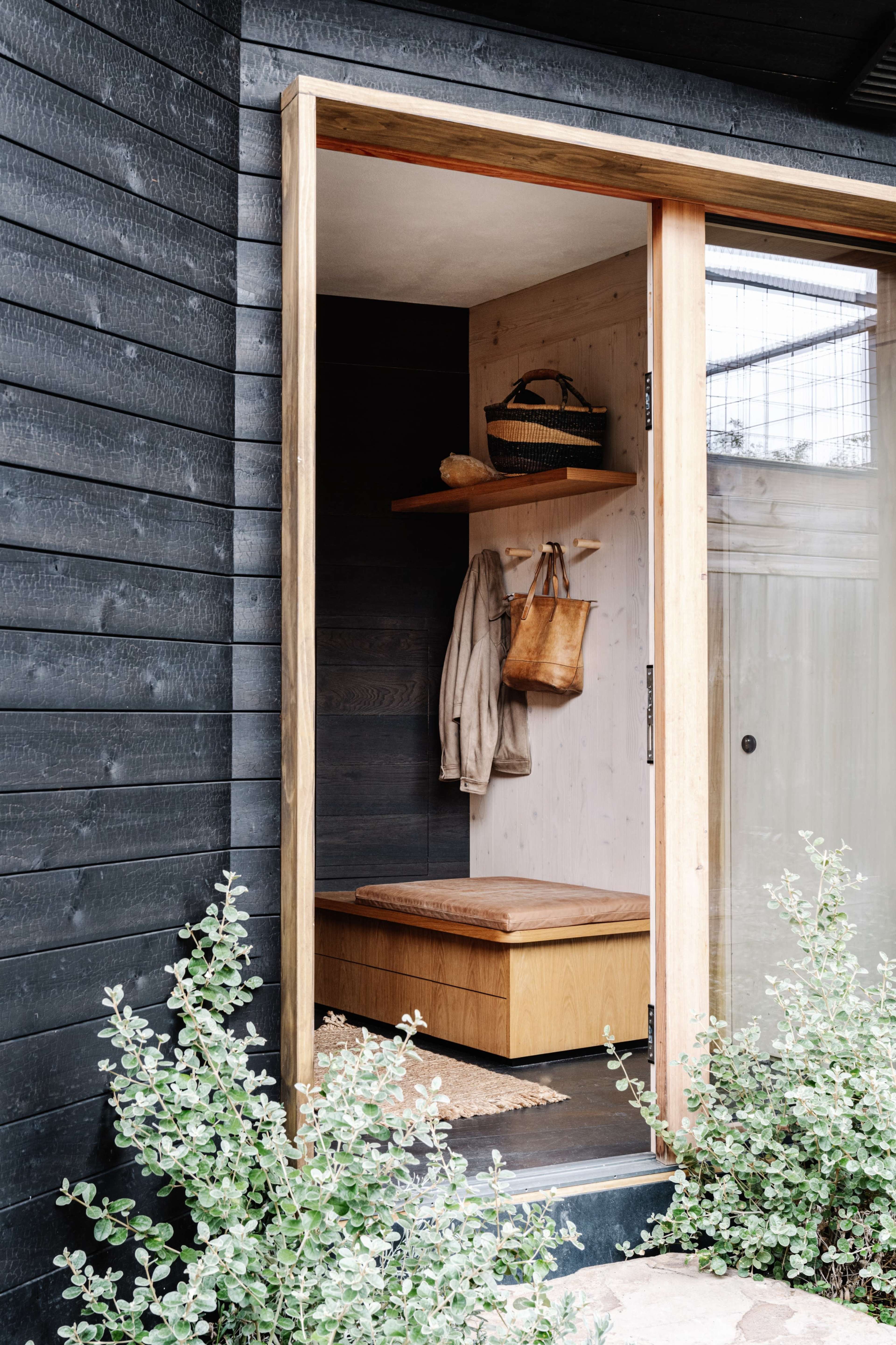 The image shows a wooden entryway with a sliding door, featuring a bench, a wall-mounted shelf with bags, and surrounding greenery.
