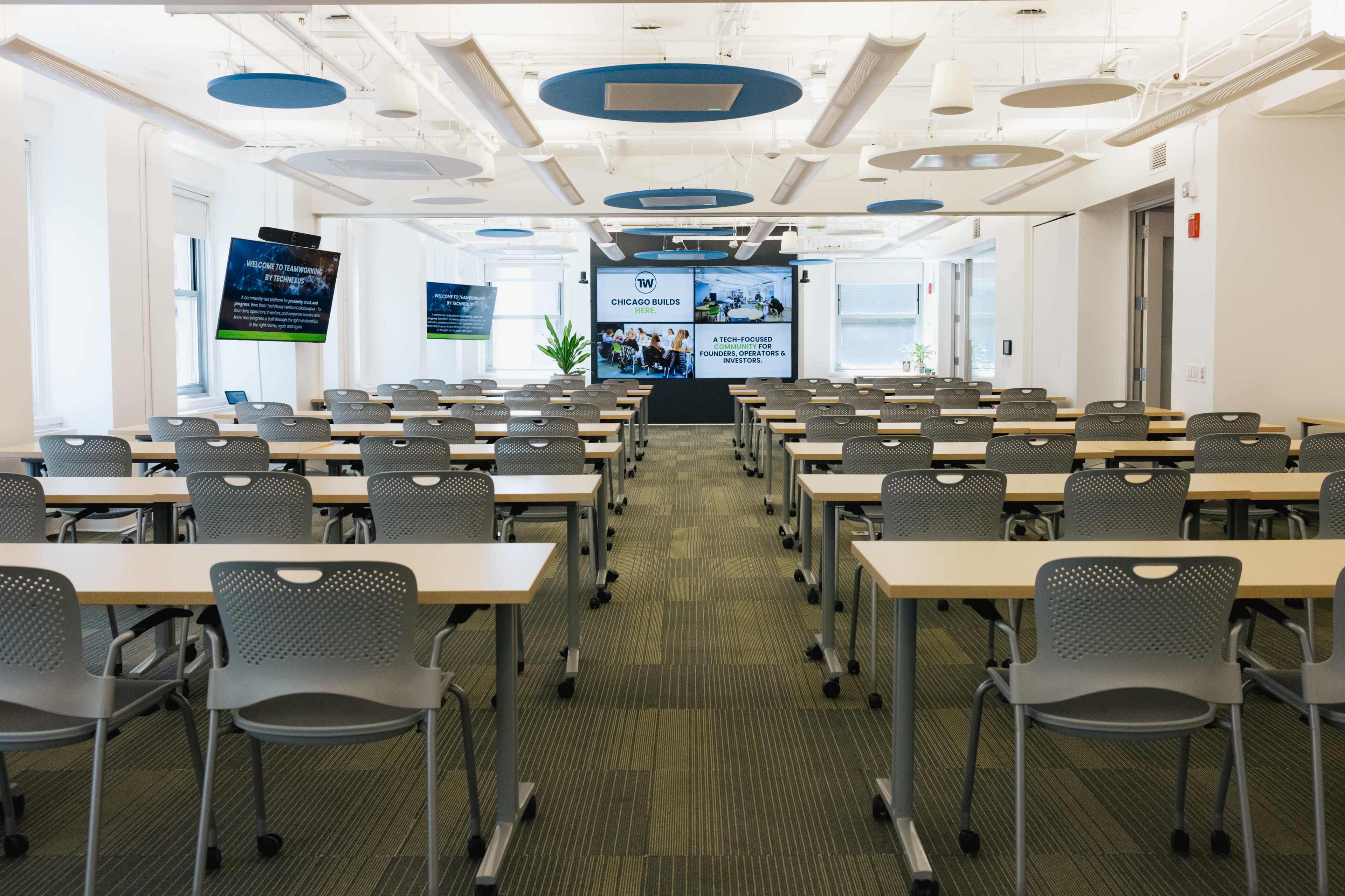 A modern classroom is set up with rows of tables and chairs, featuring a central screen displaying information in front.