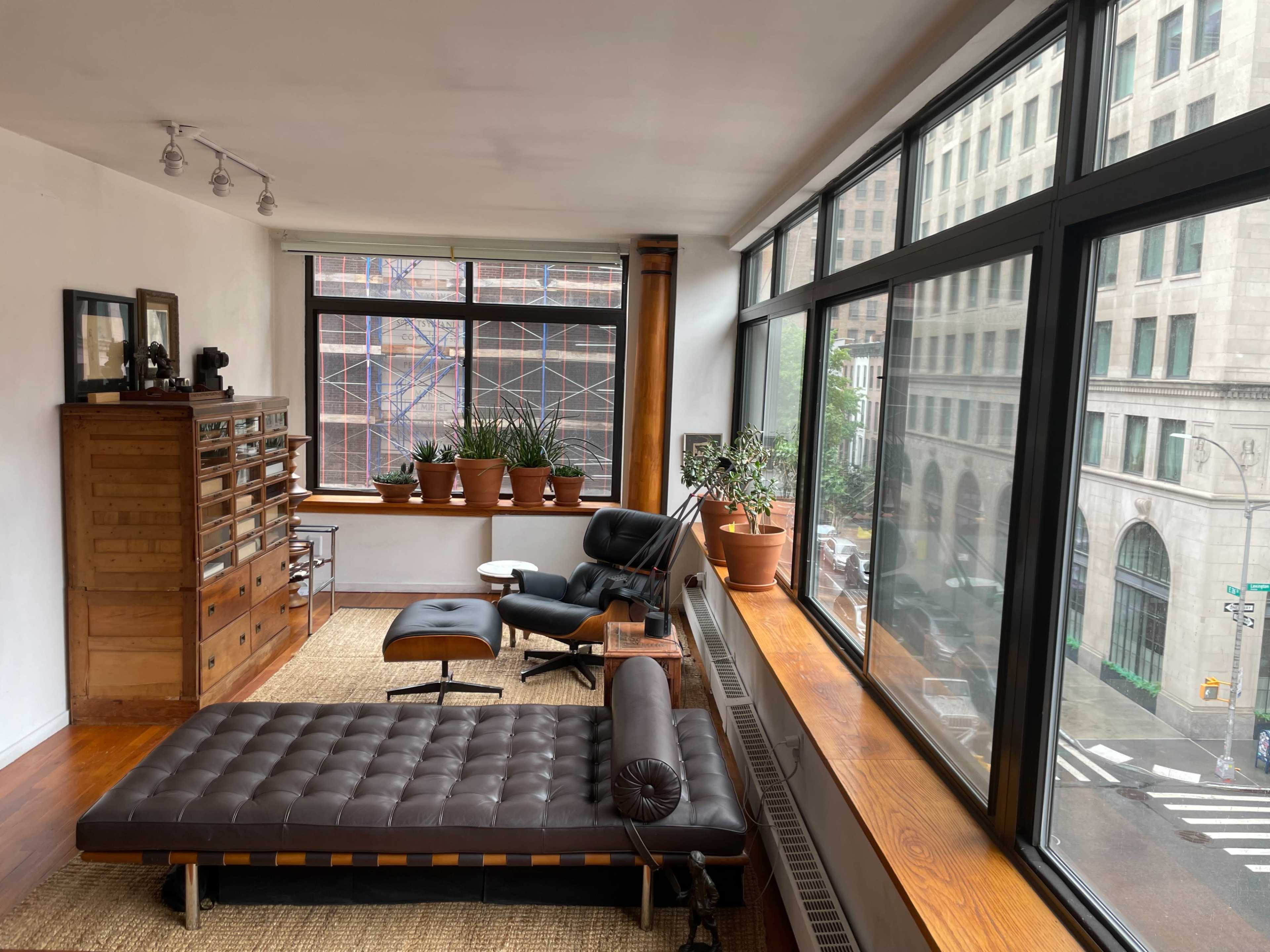 The room features a modern design with large windows, a leather lounge chair, and several potted plants, along with a wooden dresser and a distinctive chaise longue.