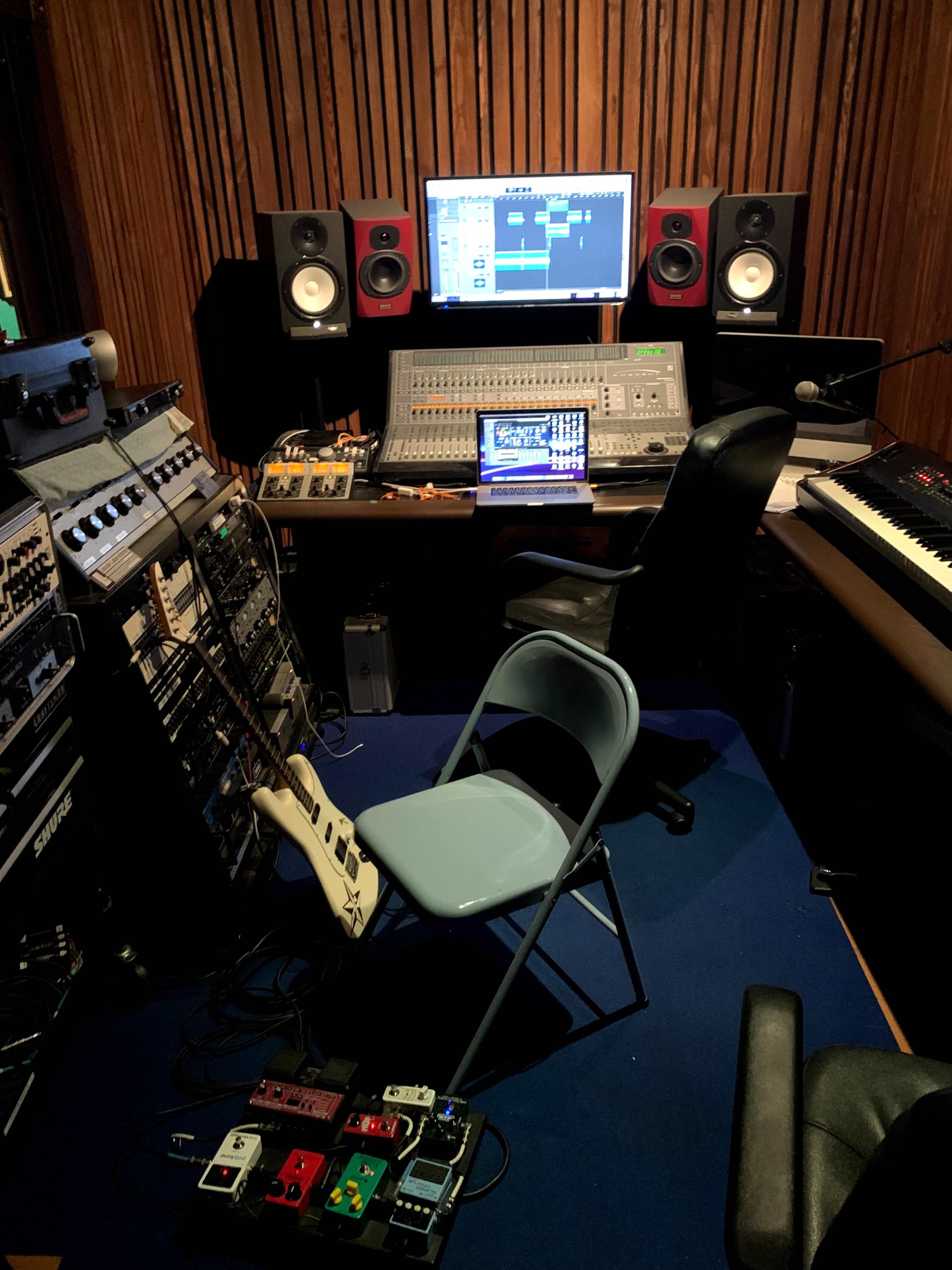 A music recording studio featuring a sound mixing console, computer screen, speakers, various equipment racks, a keyboard, and a chair.