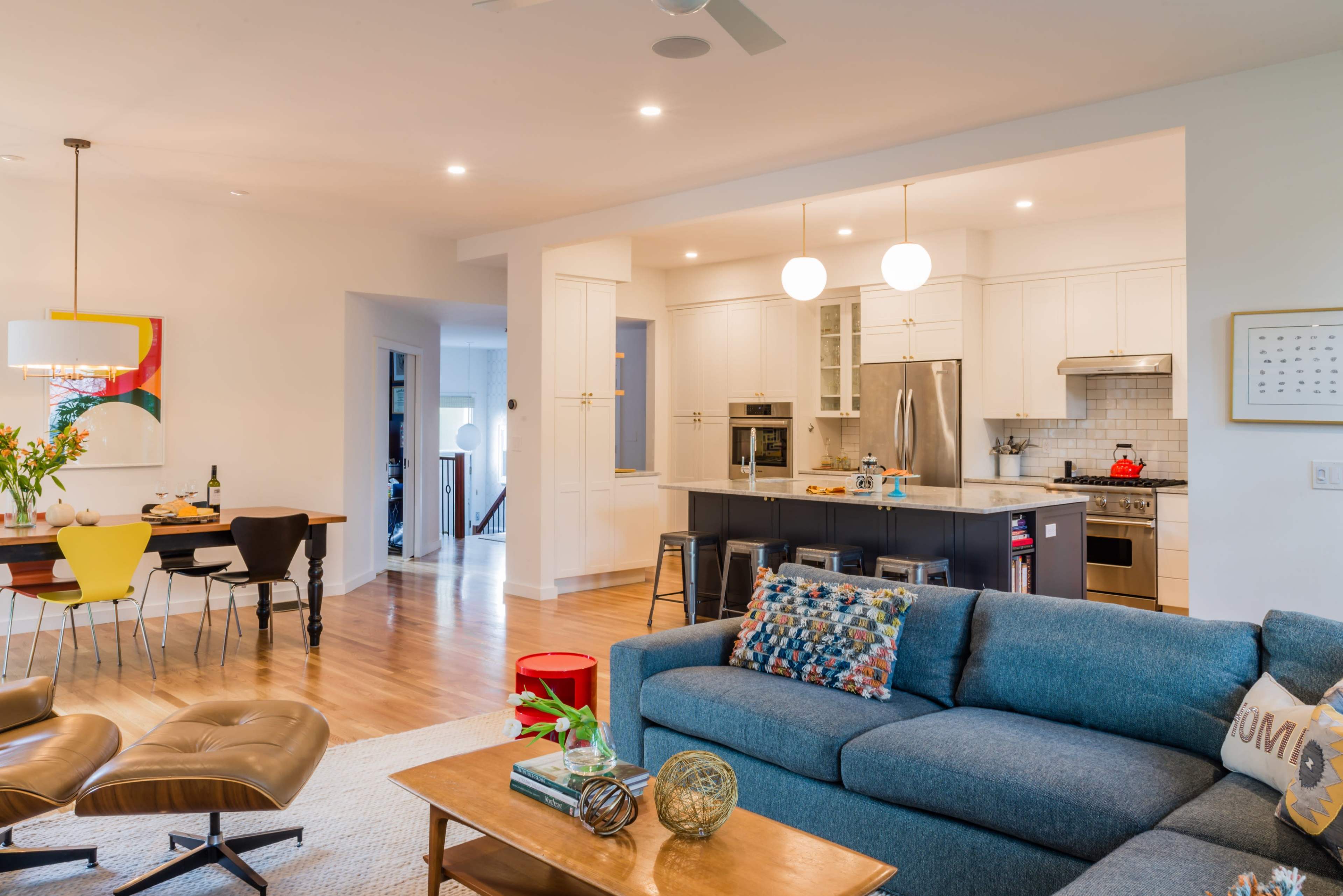 A modern living room features a blue sofa, a wooden coffee table, and an open kitchen with white cabinetry and stainless steel appliances.