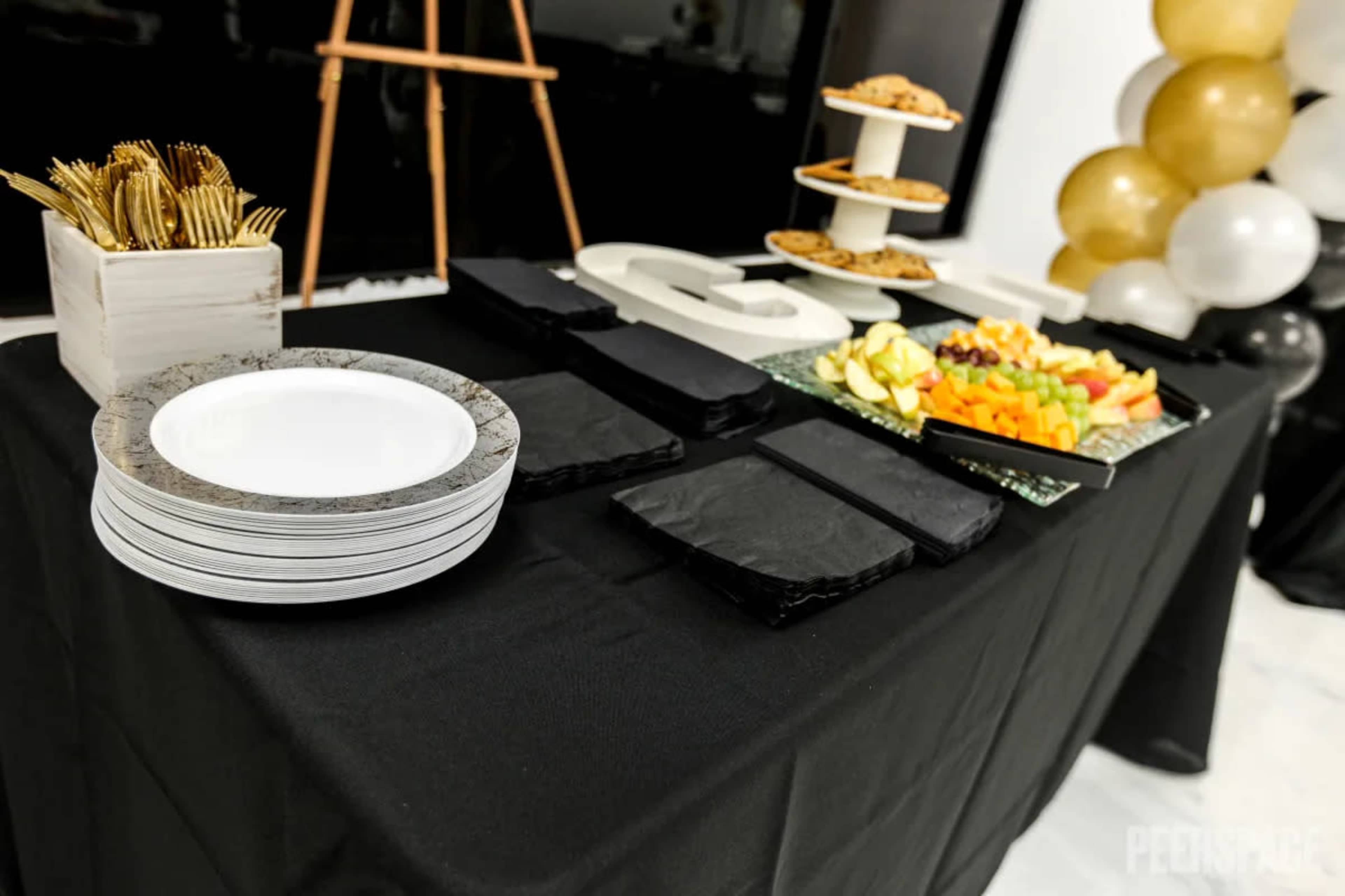 A black table displays plates, utensils, and a variety of fruits and cookies arranged on stands.