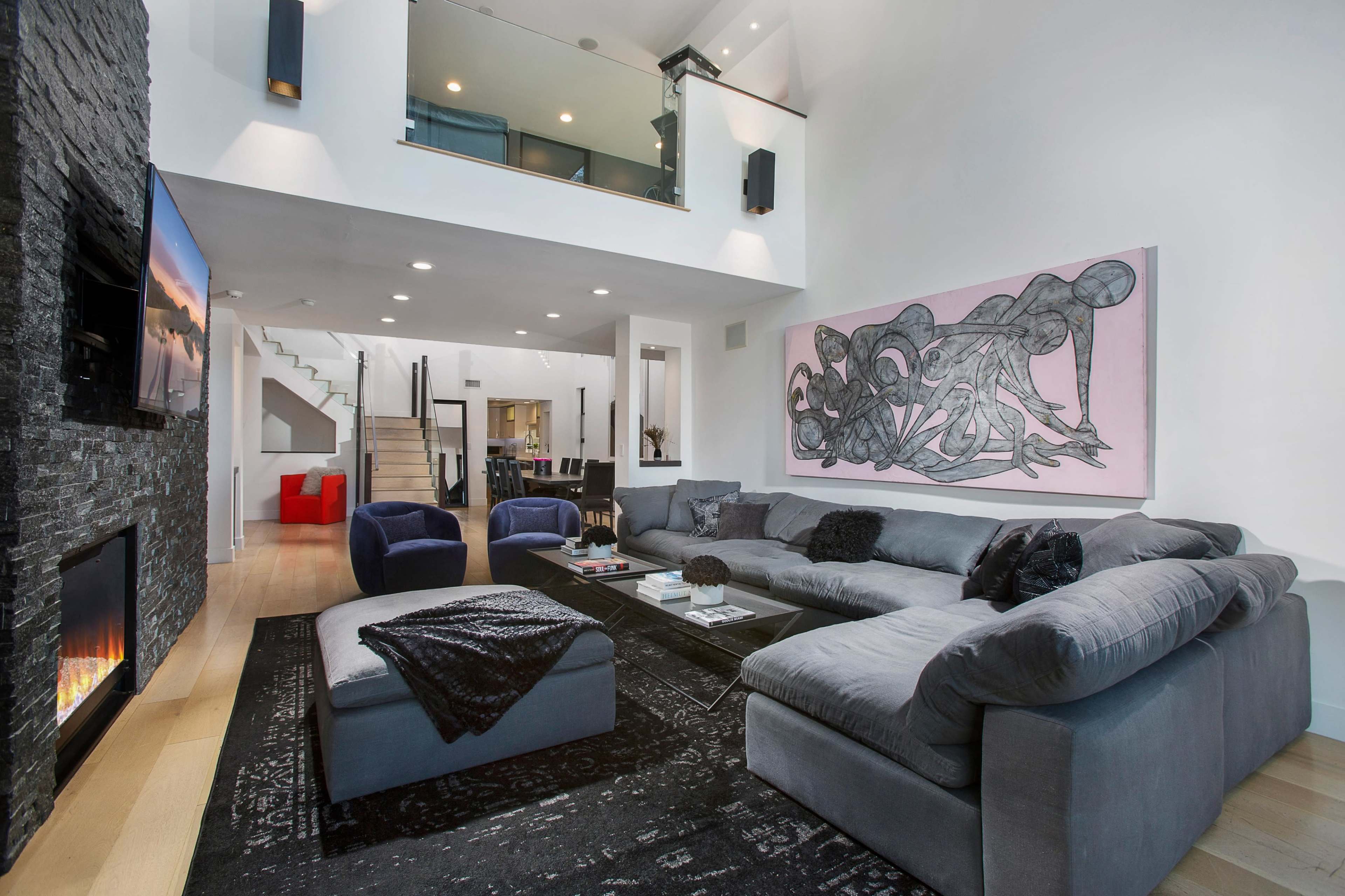 A modern living room features a large sectional sofa, a wall-mounted television, and a prominent artwork, with a staircase leading to a second floor.