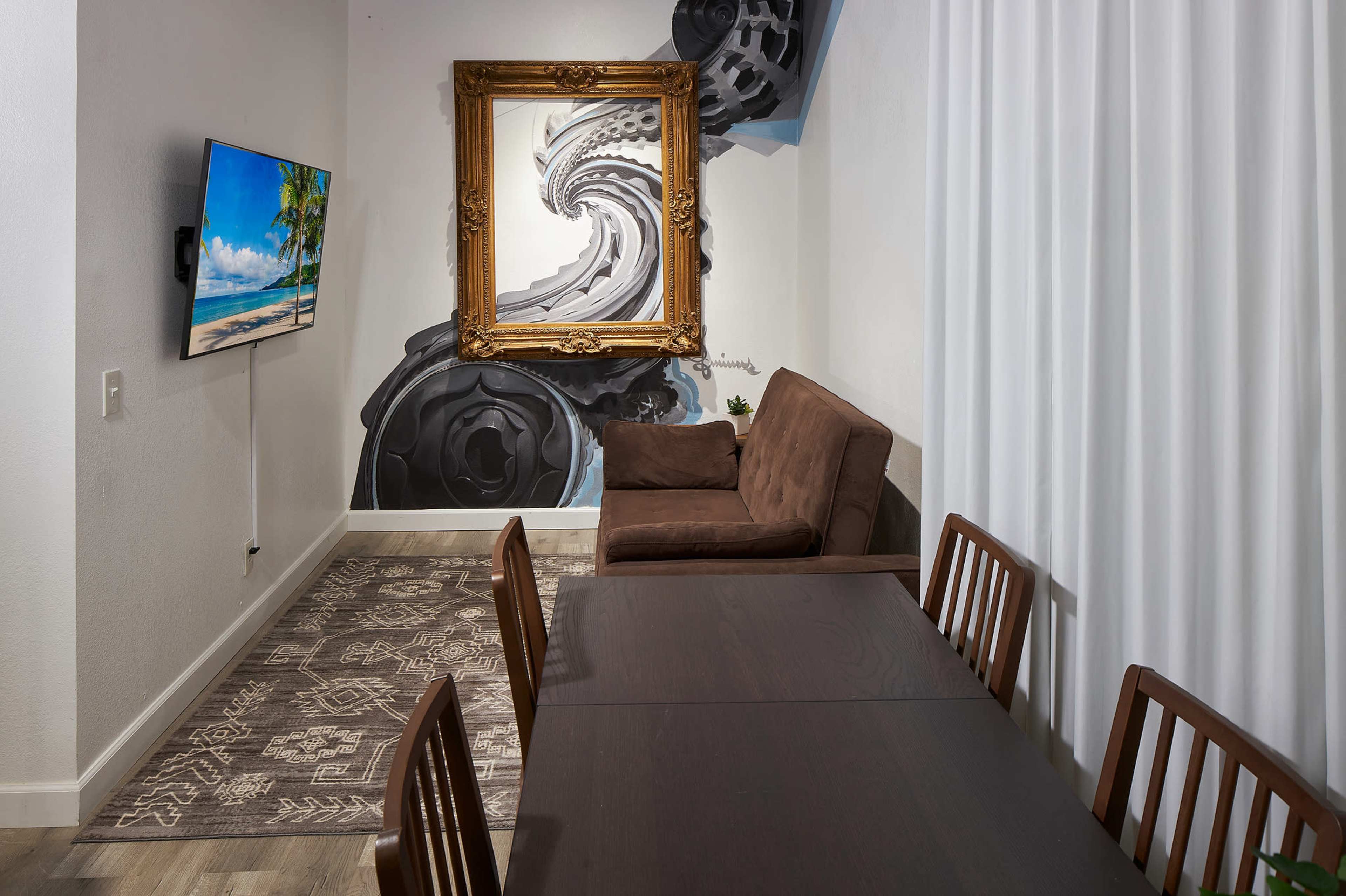 The image shows a modern living space featuring a dining table, brown couch, wall-mounted television, and an artistic mural of a spiral framed by an ornate picture frame.