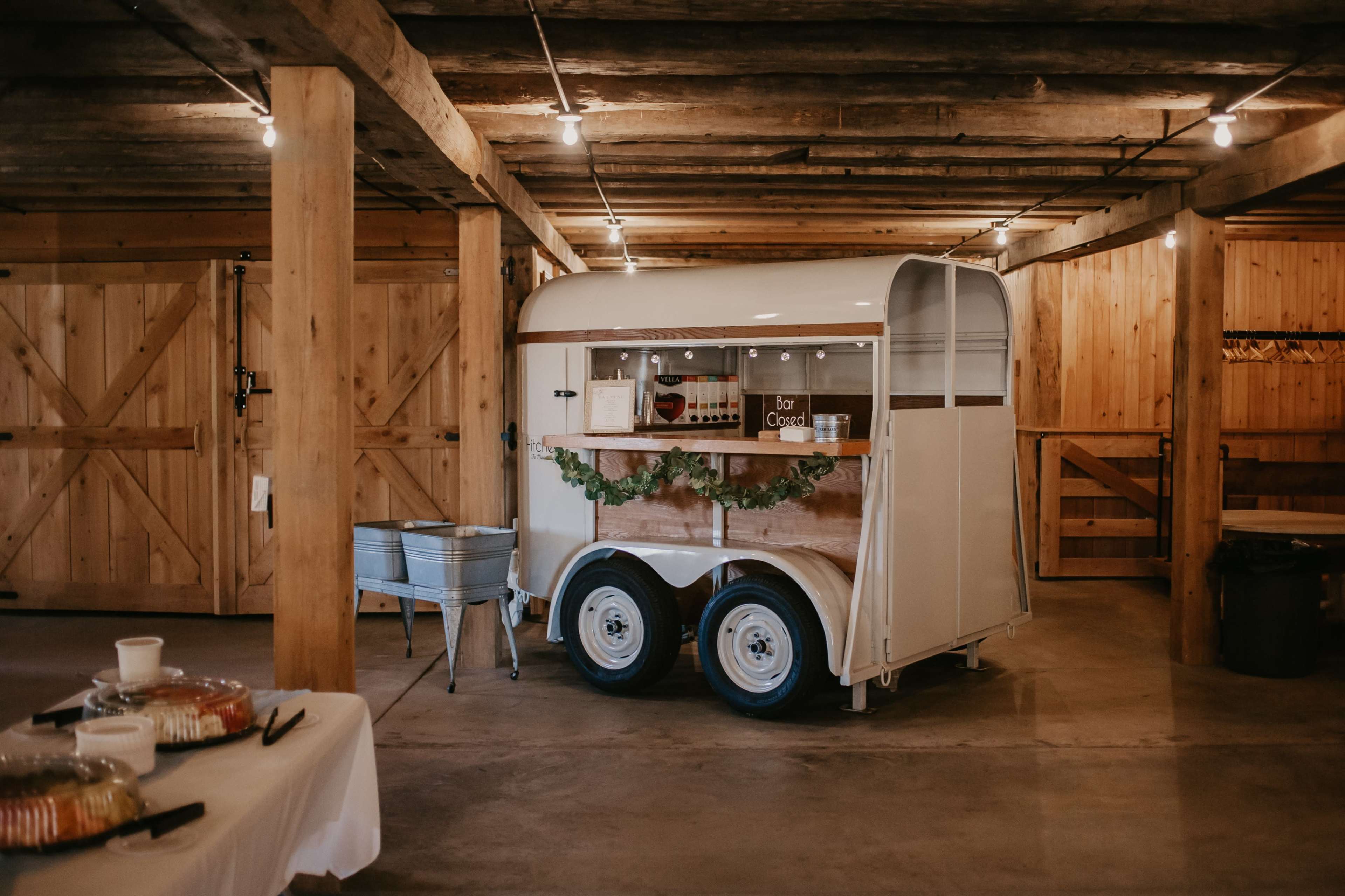 A vintage horse trailer has been converted into a mobile food stand, placed inside a rustic wooden barn with exposed beams.