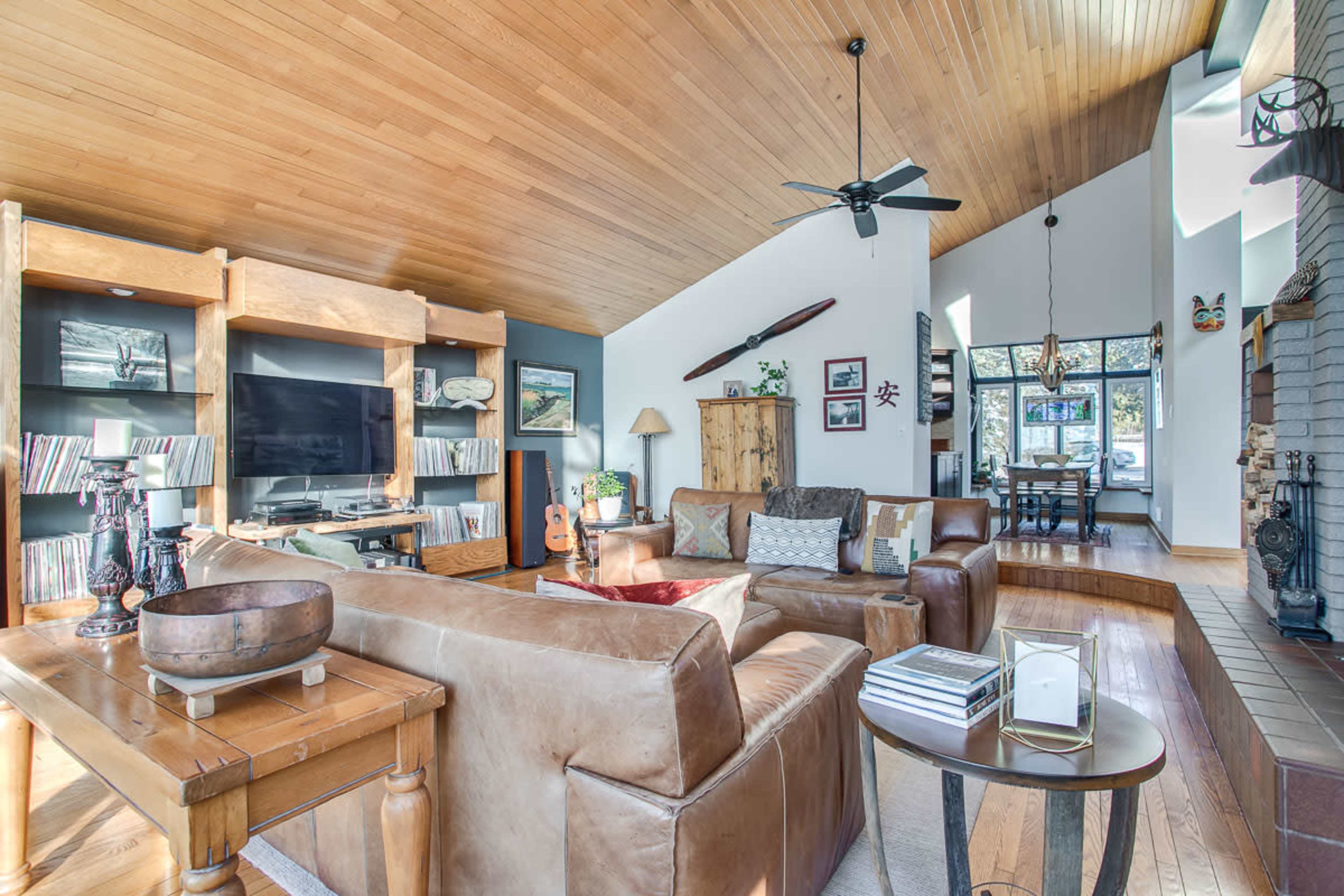 A living room with a high wooden ceiling, a large sectional sofa, a coffee table, bookshelves, and a view into a dining area.