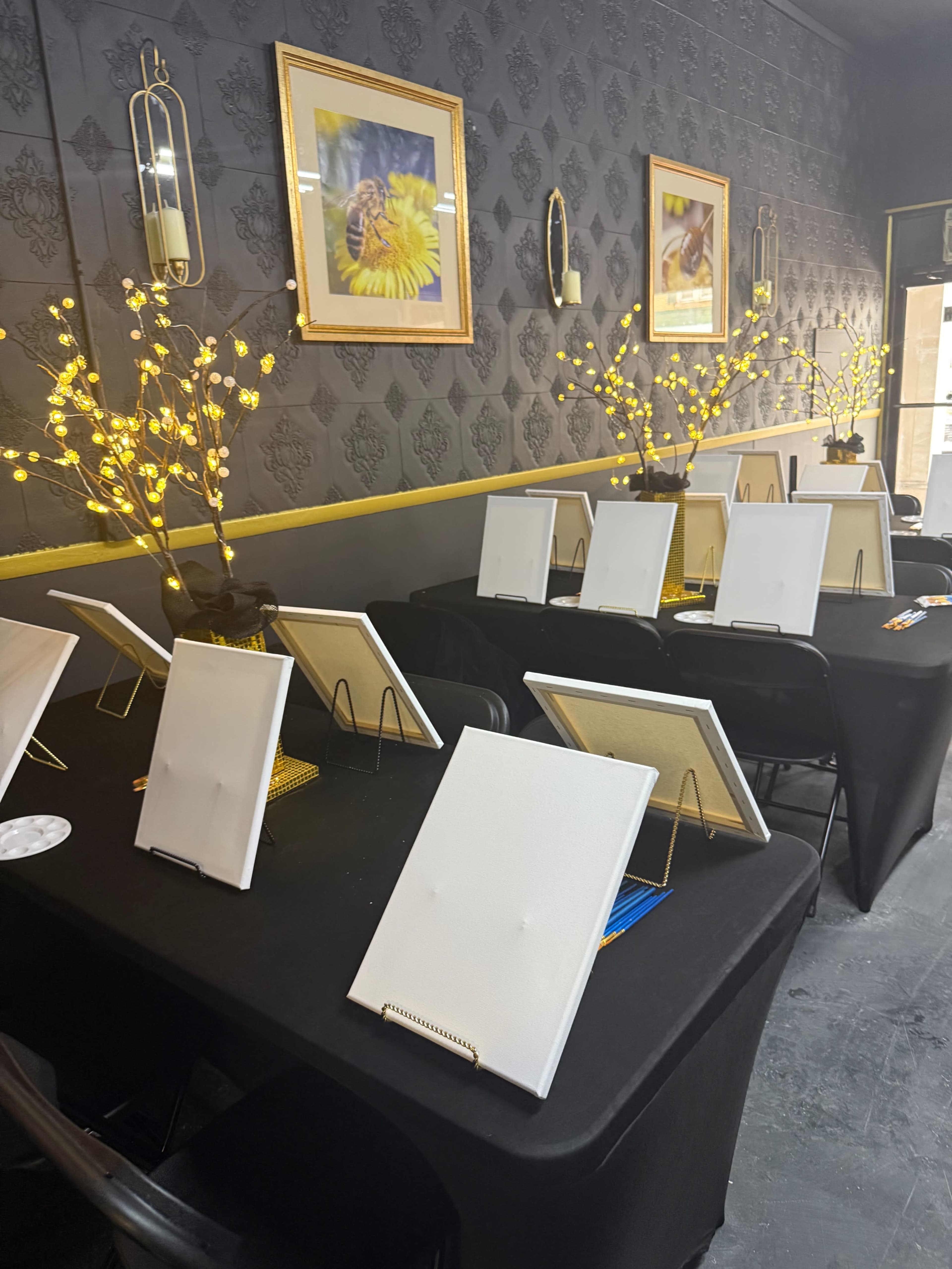 The image shows a dimly lit room with several black tables arranged, each featuring blank canvases on easels and decorative lighted branches in the background.