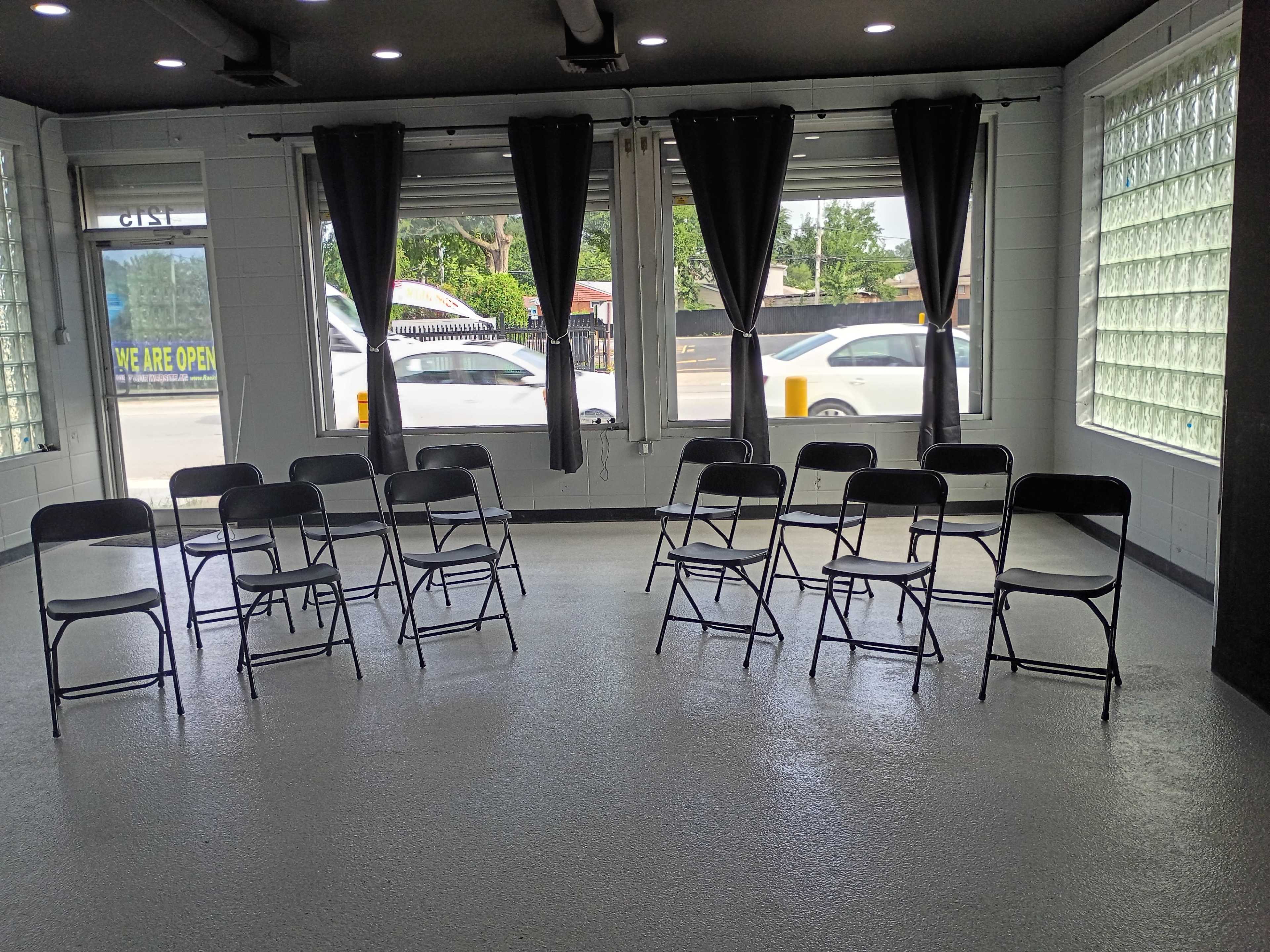A room with a smooth floor features several black chairs arranged in a circle, with large windows and light filtering through.