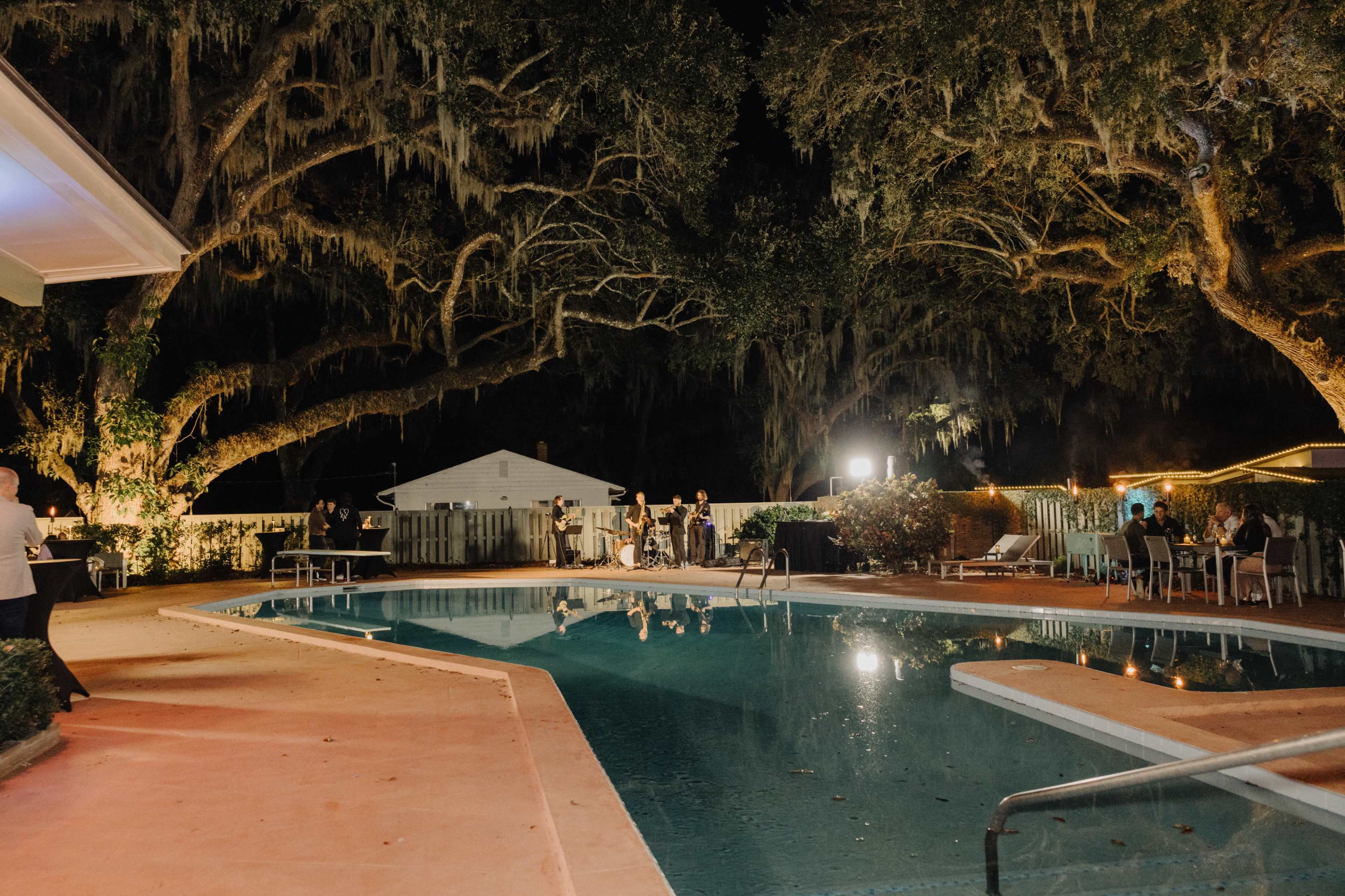 The image shows a nighttime scene of a pool area surrounded by large trees with Spanish moss, featuring several people socializing near the water's edge.