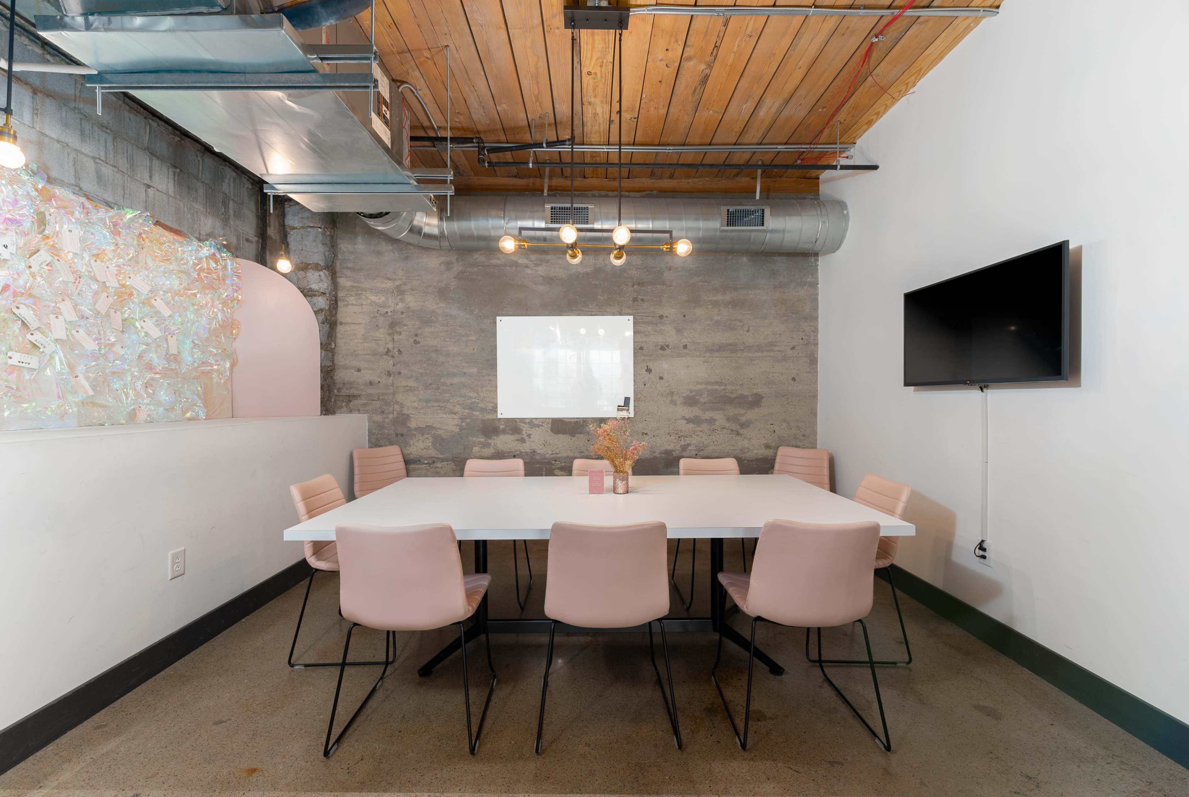 A modern conference room features a long white table surrounded by pink chairs, with exposed wooden beams and a large screen on the wall.