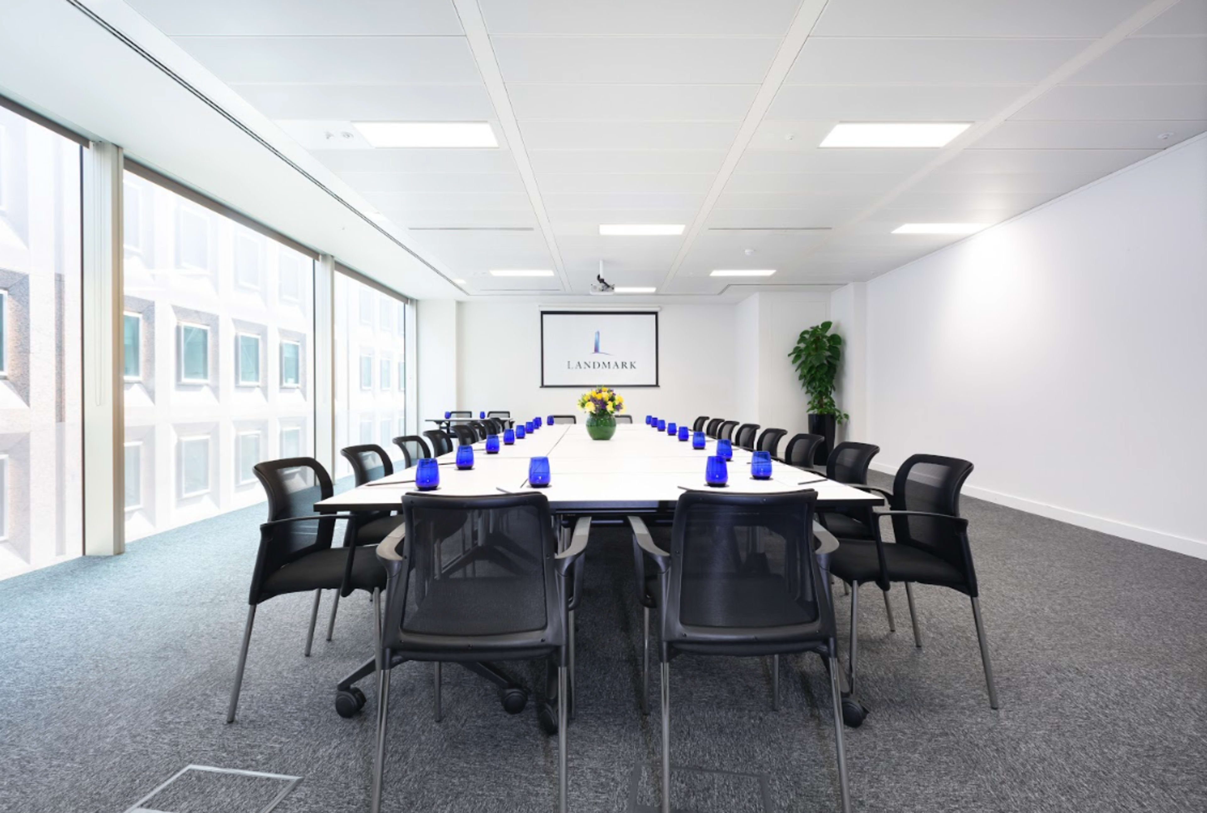 A modern conference room features a long table surrounded by black chairs, with a large screen displaying a logo and blue glass vases arranged along the center.