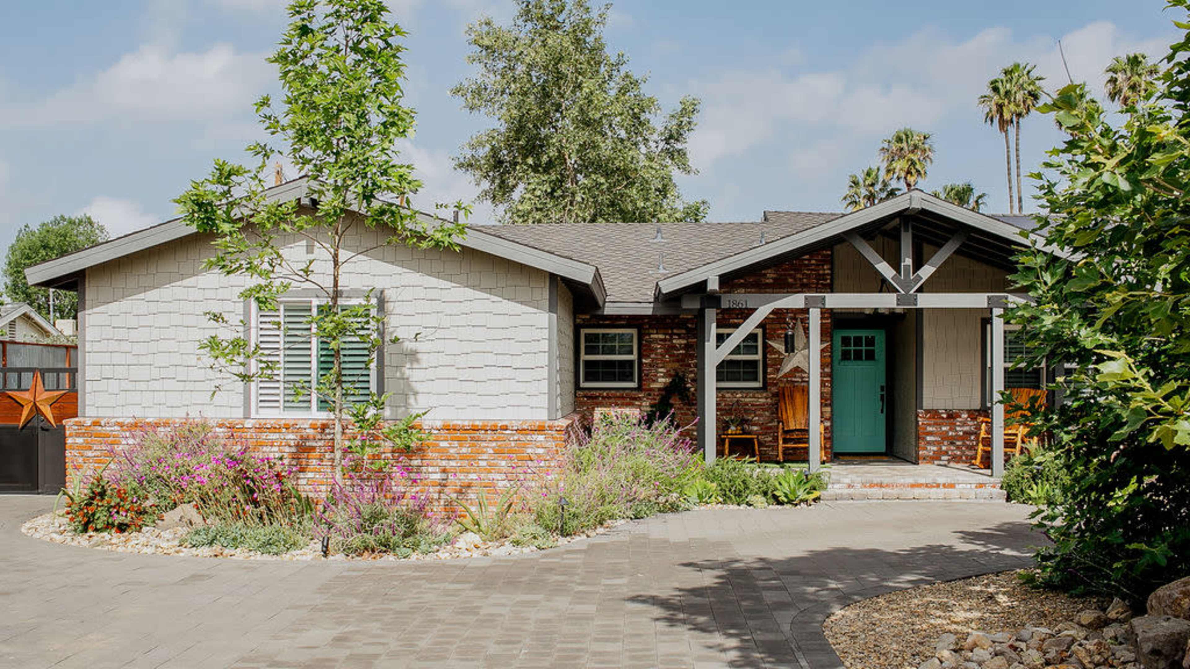 The image shows a single-story house with a combination of brick and stucco exteriors, surrounded by landscaped gardens and a paved driveway.