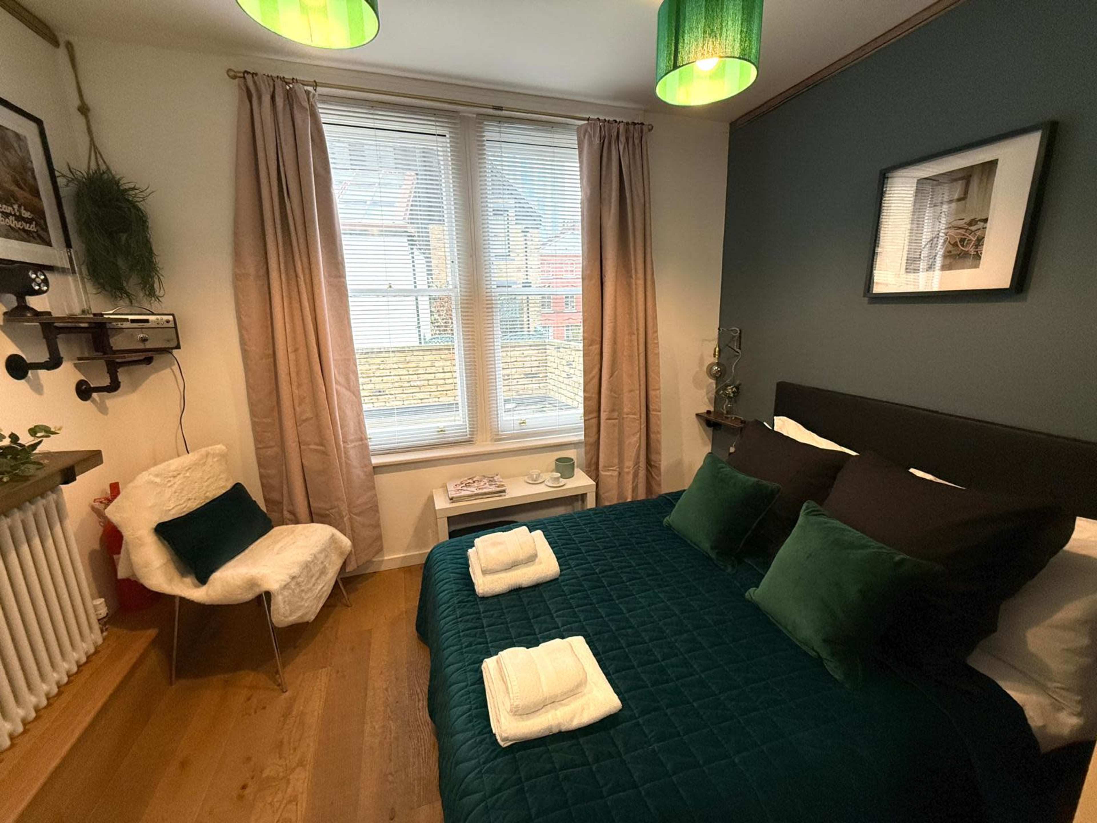 The image shows a cozy bedroom featuring a green bedspread, two pillows, a small white chair, and large windows with natural light.