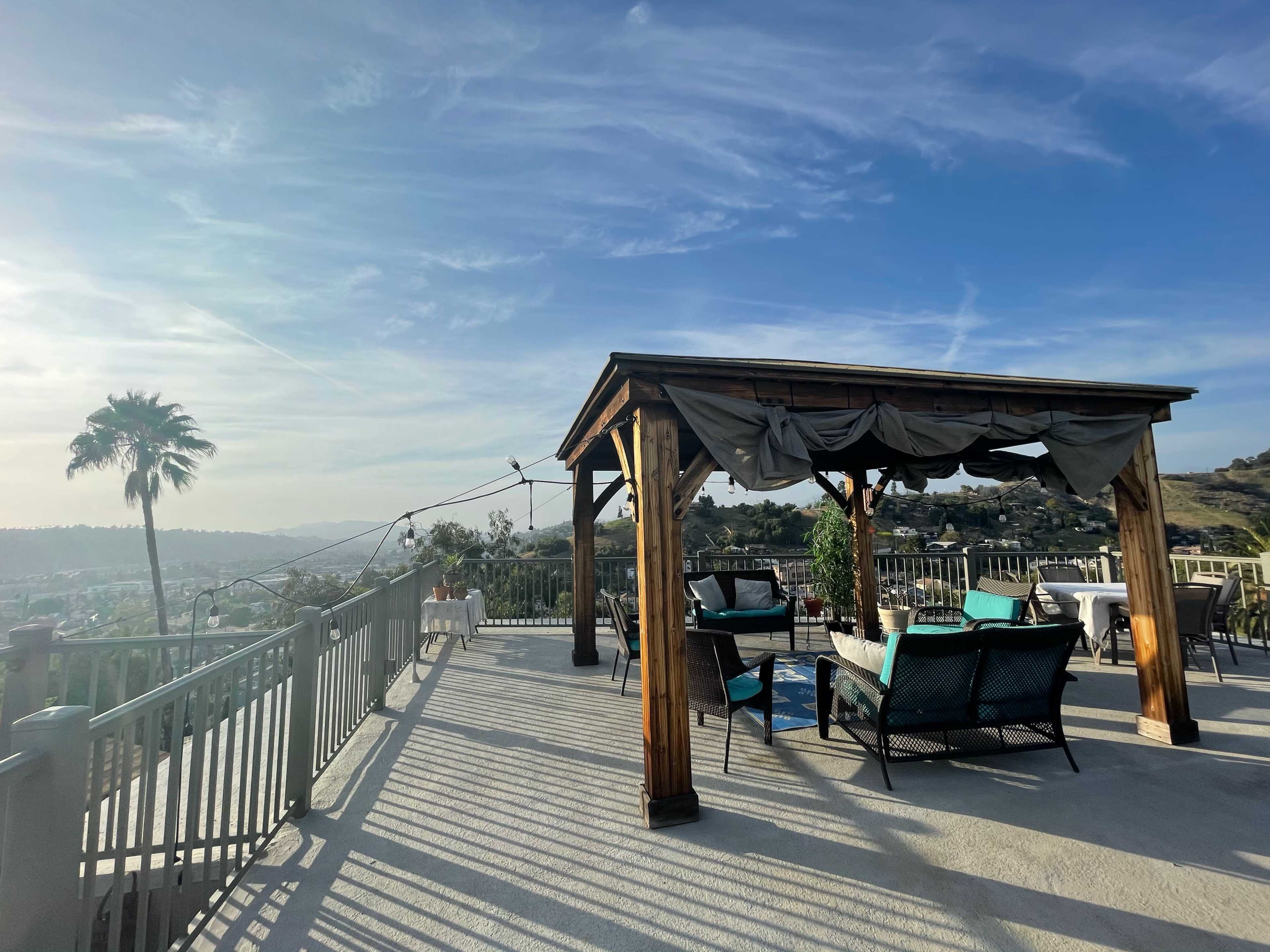 A wooden pergola with seating overlooks a valley under a clear sky.