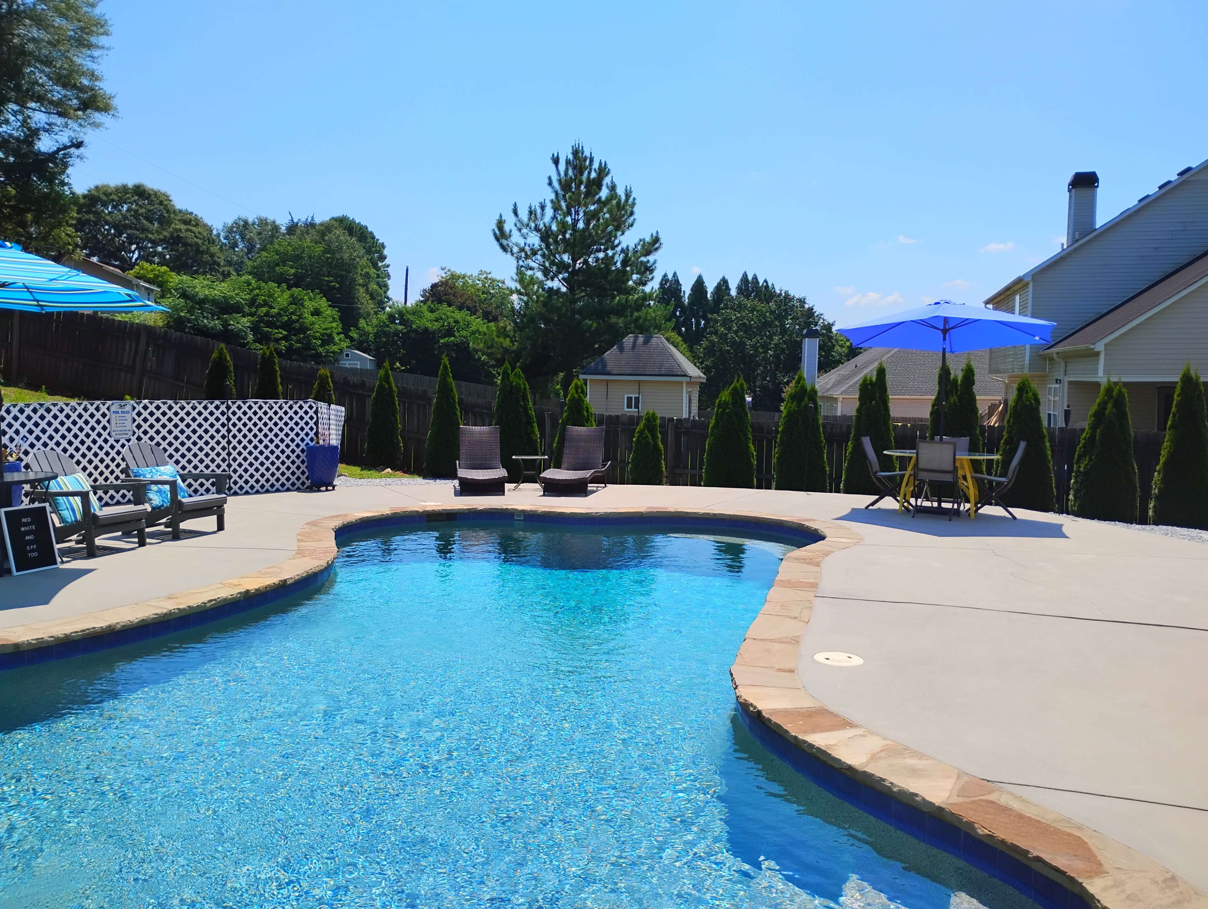 The image shows a swimming pool surrounded by lounge chairs and tall evergreen shrubs under a clear blue sky.