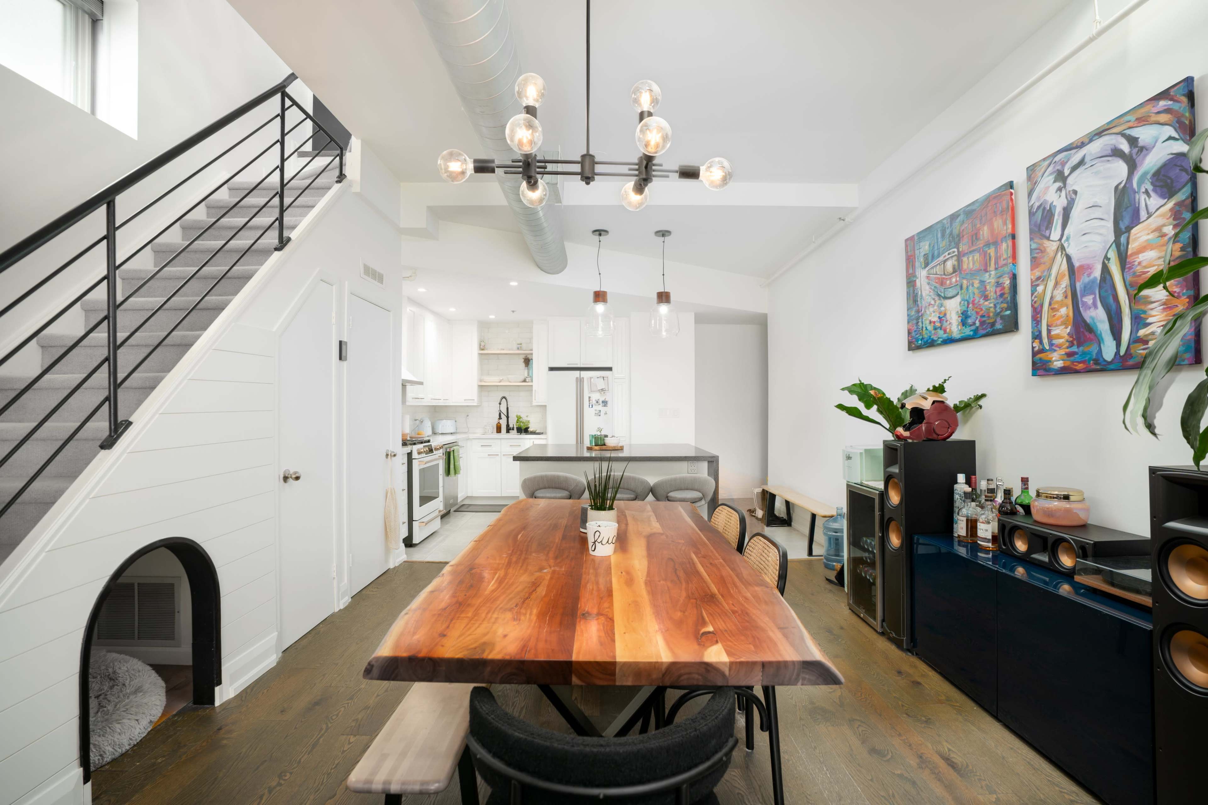 A modern dining area with a wooden table, a staircase leading to an upper level, and an open kitchen in the background featuring white cabinets and appliances.