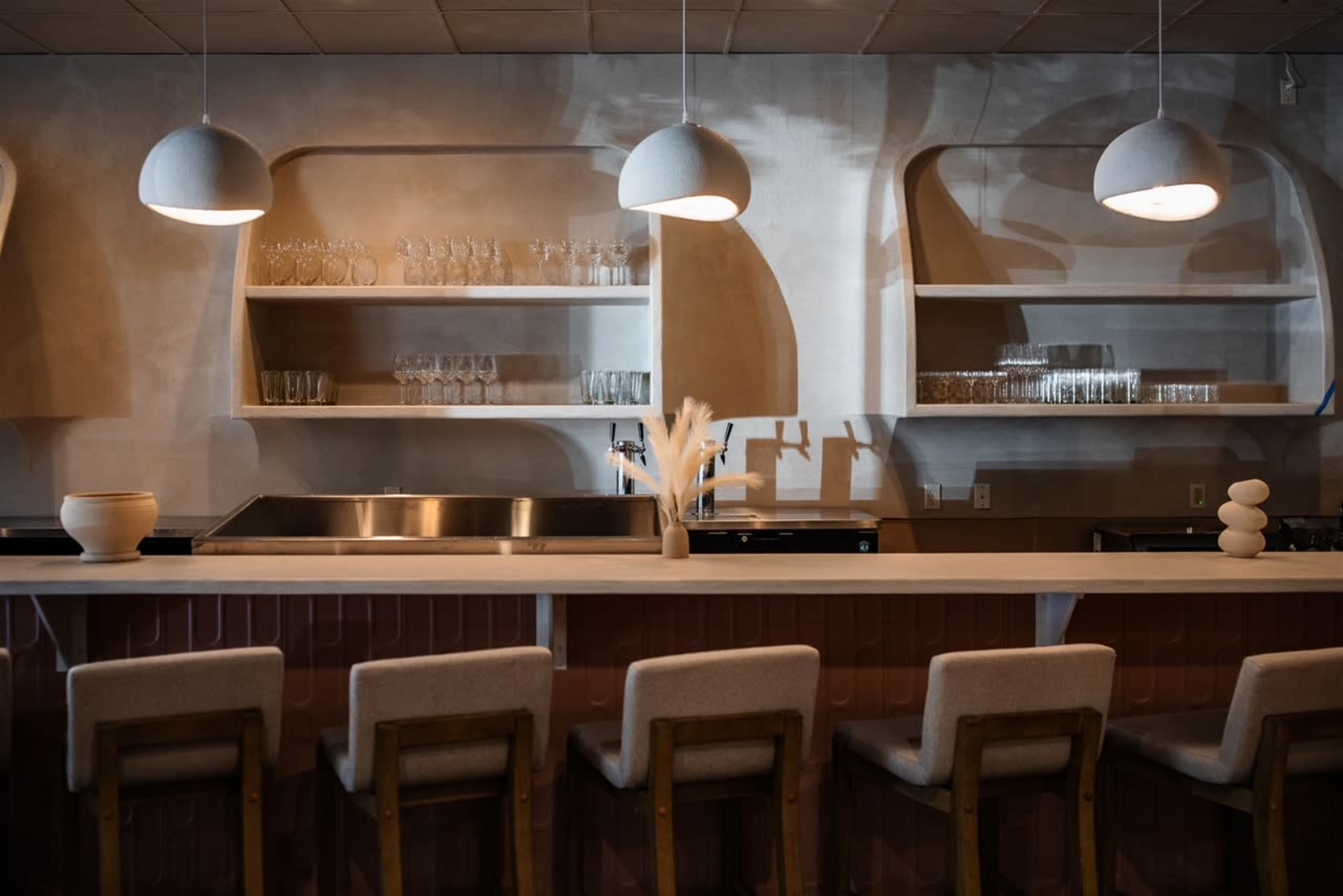 The image shows a minimalistic bar area with a long countertop, shelving displaying glassware, and soft, modern lighting.