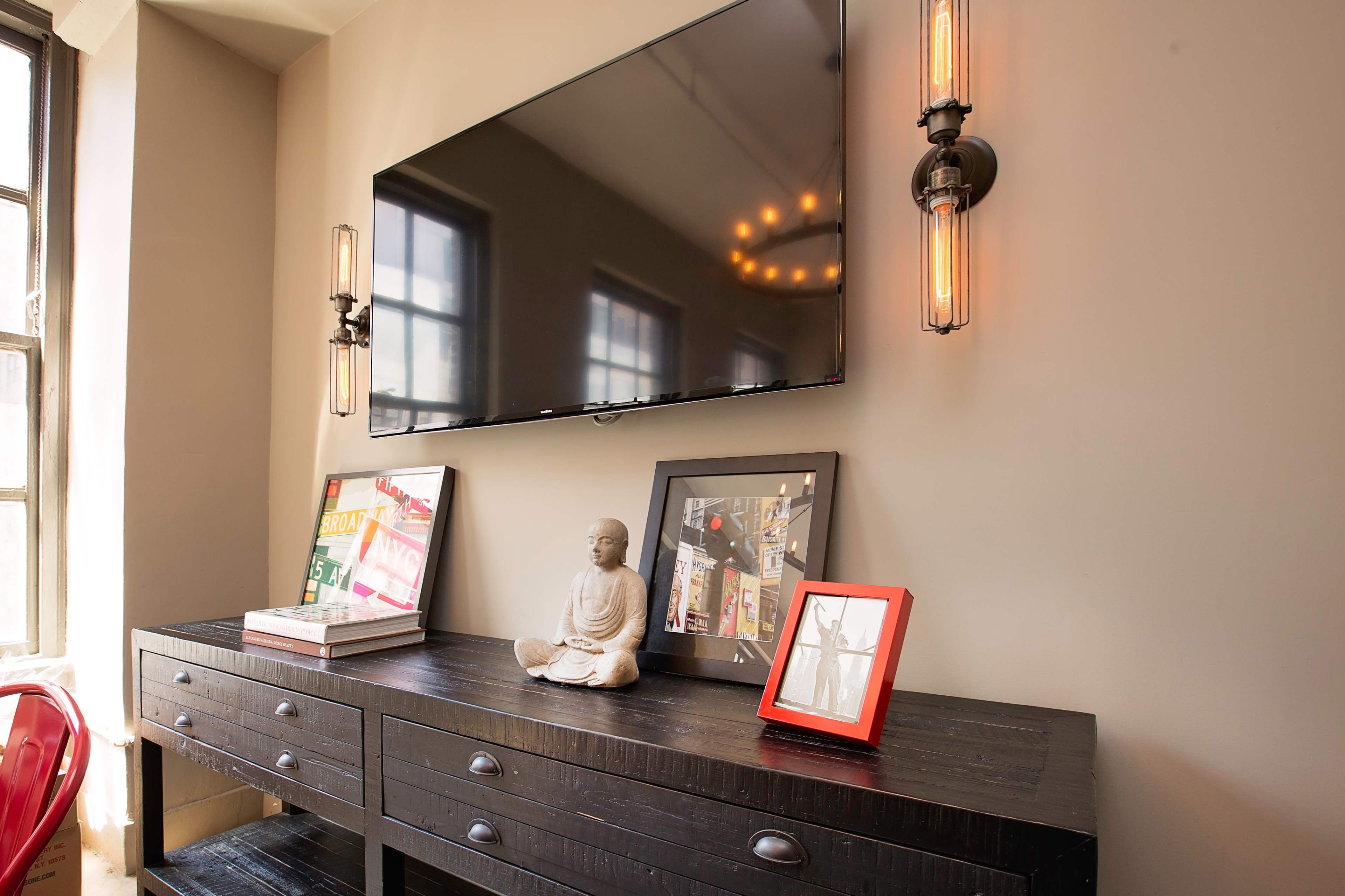 A wall-mounted television is situated above a dark wooden console, which displays a statue and framed photographs against a backdrop of windows.