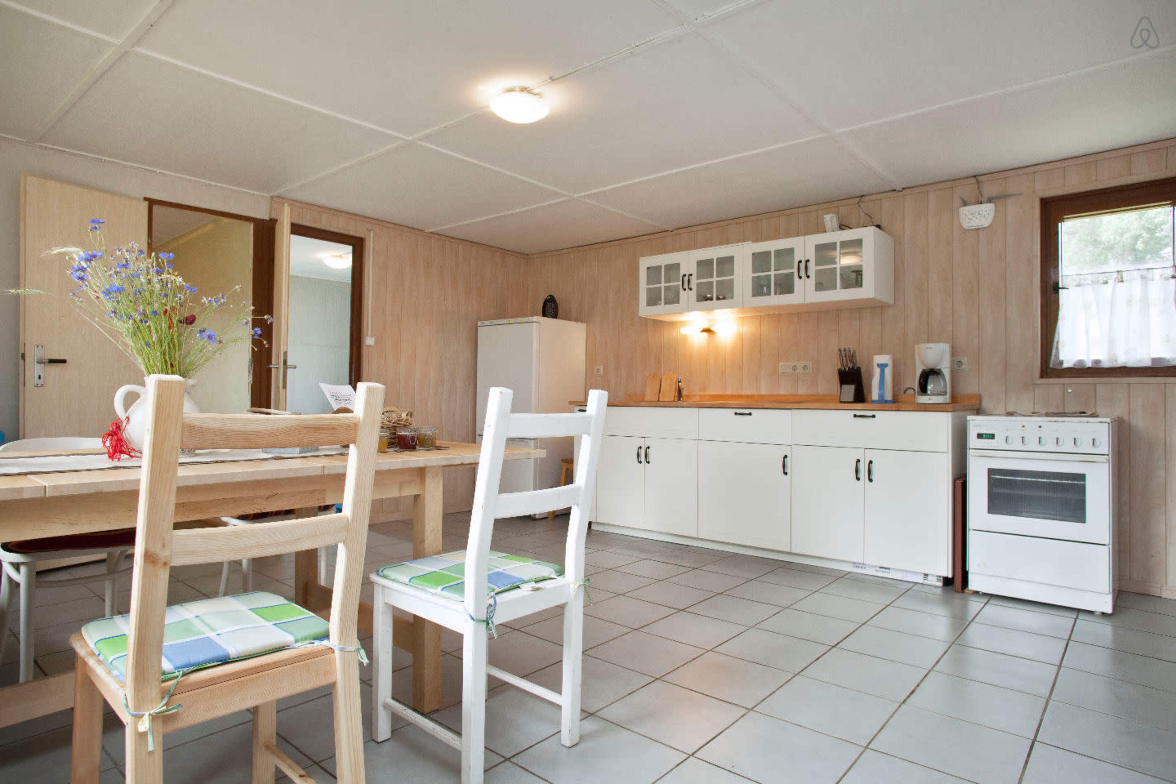 The image shows a simple kitchen area with a wooden dining table, white cabinets, a stove, and a refrigerator, all set against a wall of light wood paneling.